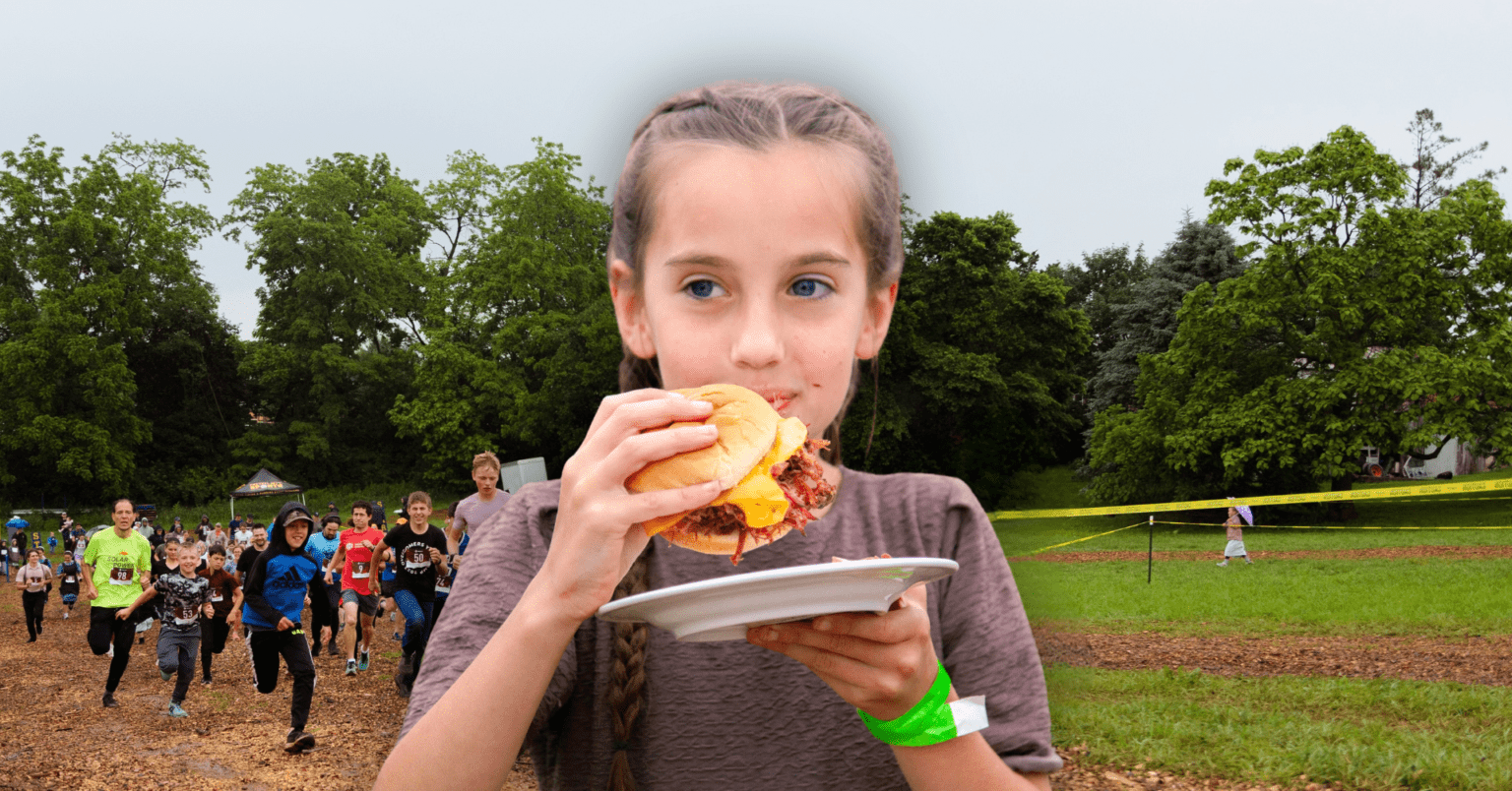 Girl enjoying a burger on a plate at a community running event, with participants running in the background surrounded by trees and greenery.