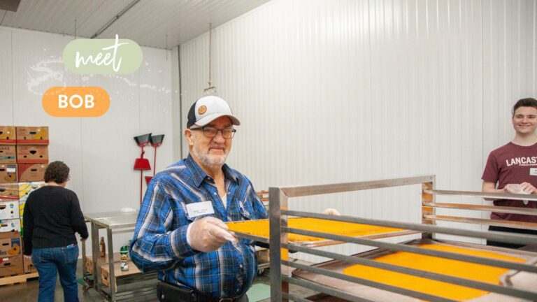 Senior volunteer Bob working in a food production facility, handling trays of vibrant orange food product, while a young man smiles in the background. The image captures a collaborative and community-focused environment.