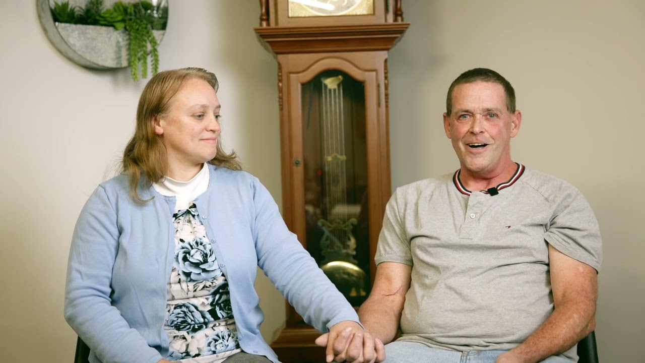 Smiling couple sitting and holding hands in cozy room with clock in background.