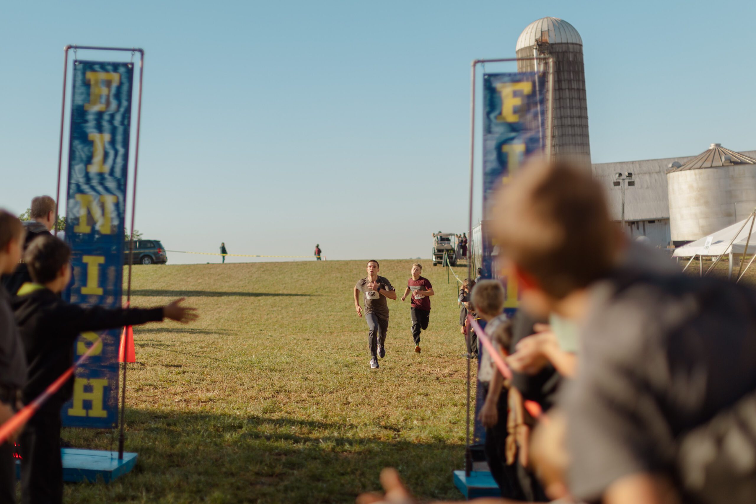 Boys sprinting toward the finish line at a running event, surrounded by cheering spectators, with a barn and silo in the background under a clear blue sky.