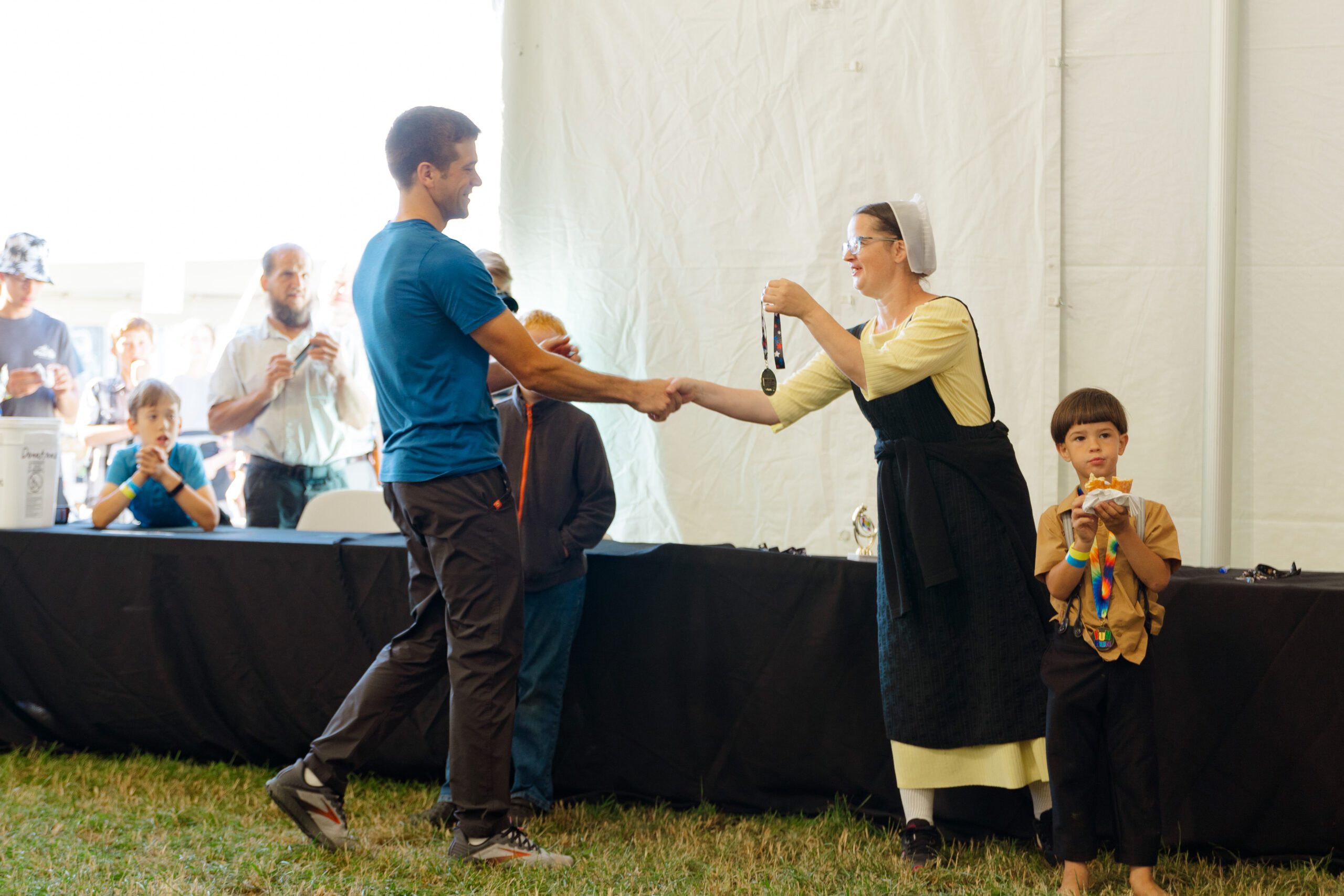 A medal ceremony taking place inside a tent, featuring a man in a blue shirt shaking hands with a woman in traditional attire who is presenting a medal. Children observe the event, with one boy holding a snack and a colorful necklace. The atmosphere is festive and celebratory.