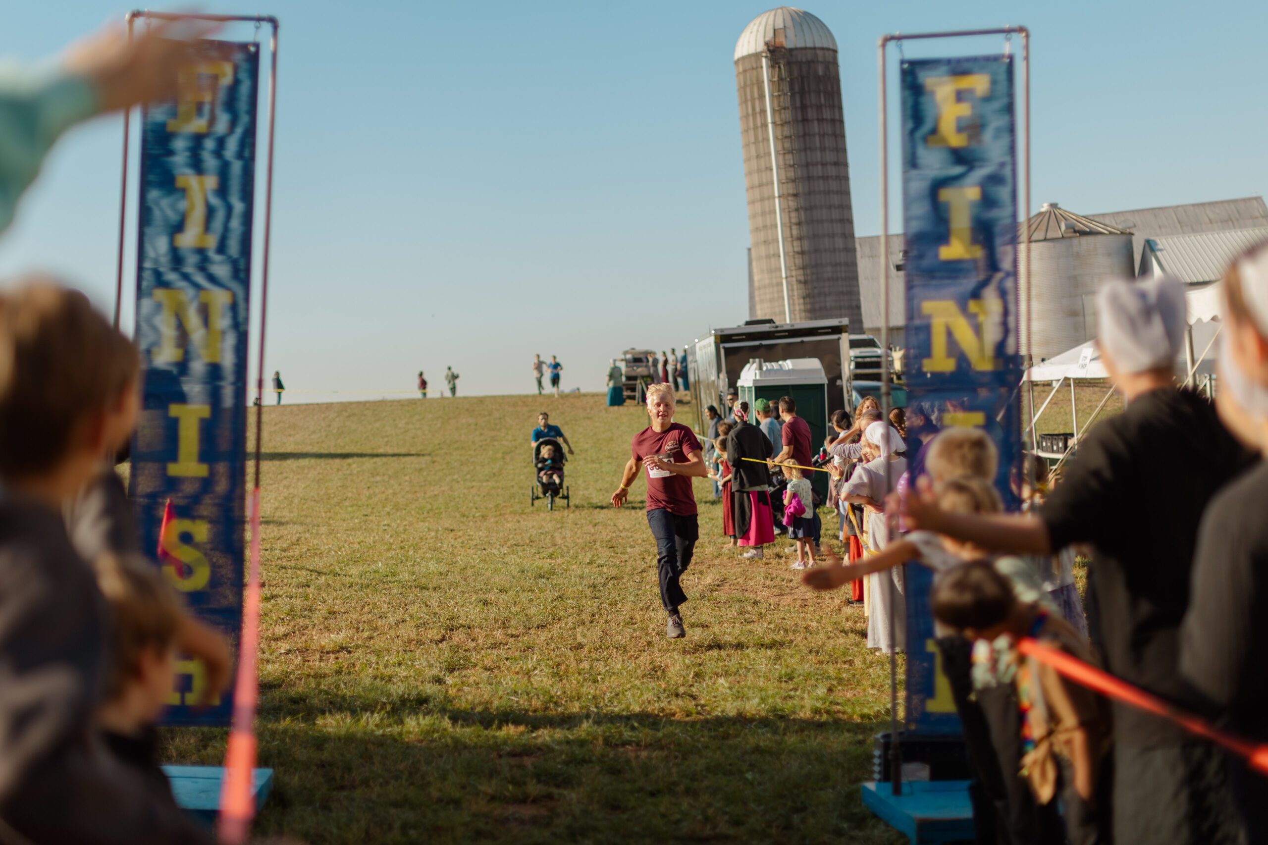 A young boy in a maroon shirt sprints toward the finish line at an outdoor event, surrounded by a crowd of spectators. A red ribbon marks the finish area, with banners labeled "FINISH" on either side. In the background, a farm setting with a silo and people enjoying the festivities is visible under a clear blue sky.