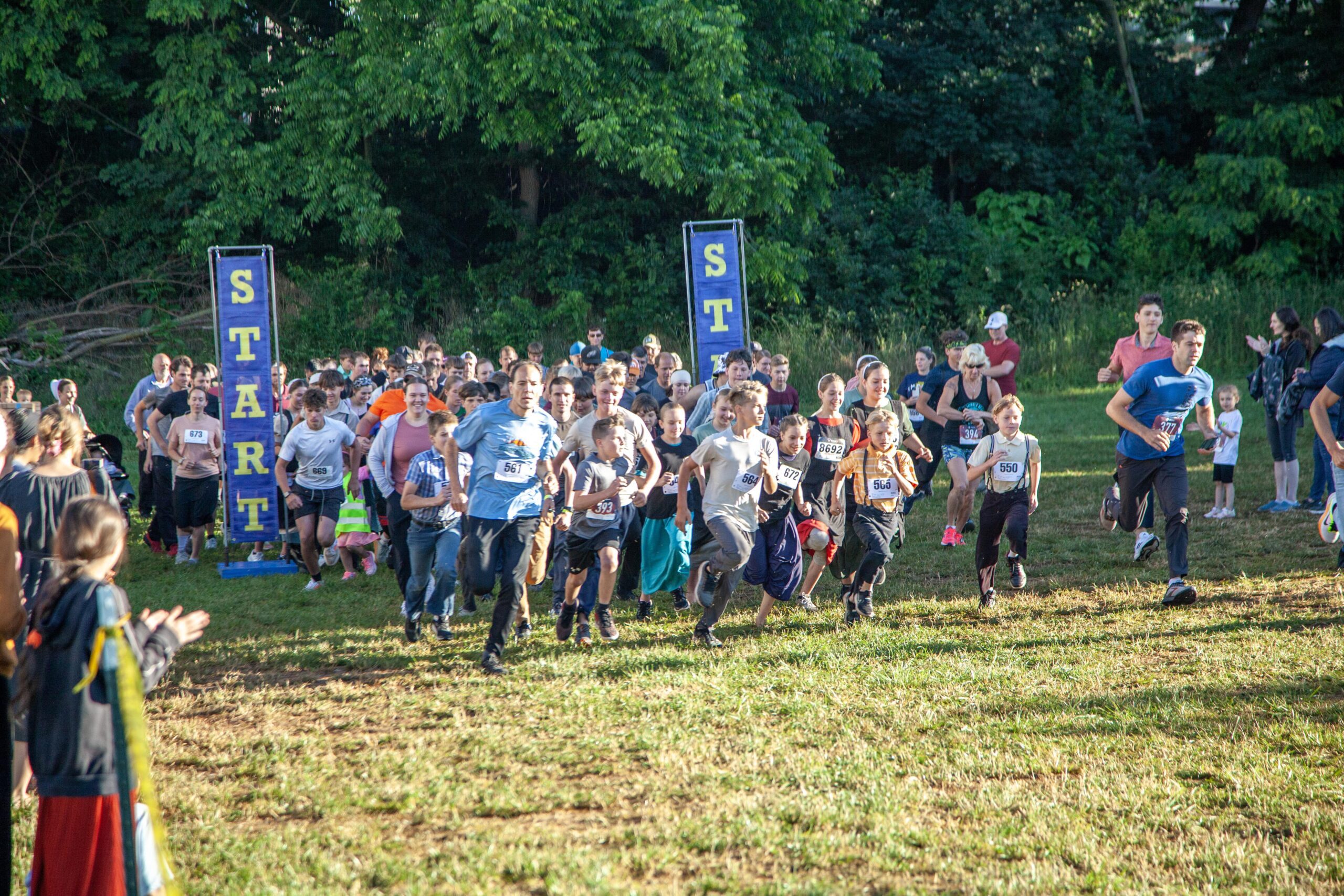 A large group of diverse participants, including adults and children, enthusiastically starting a running race marked by a "START" banner on a grassy field surrounded by trees.