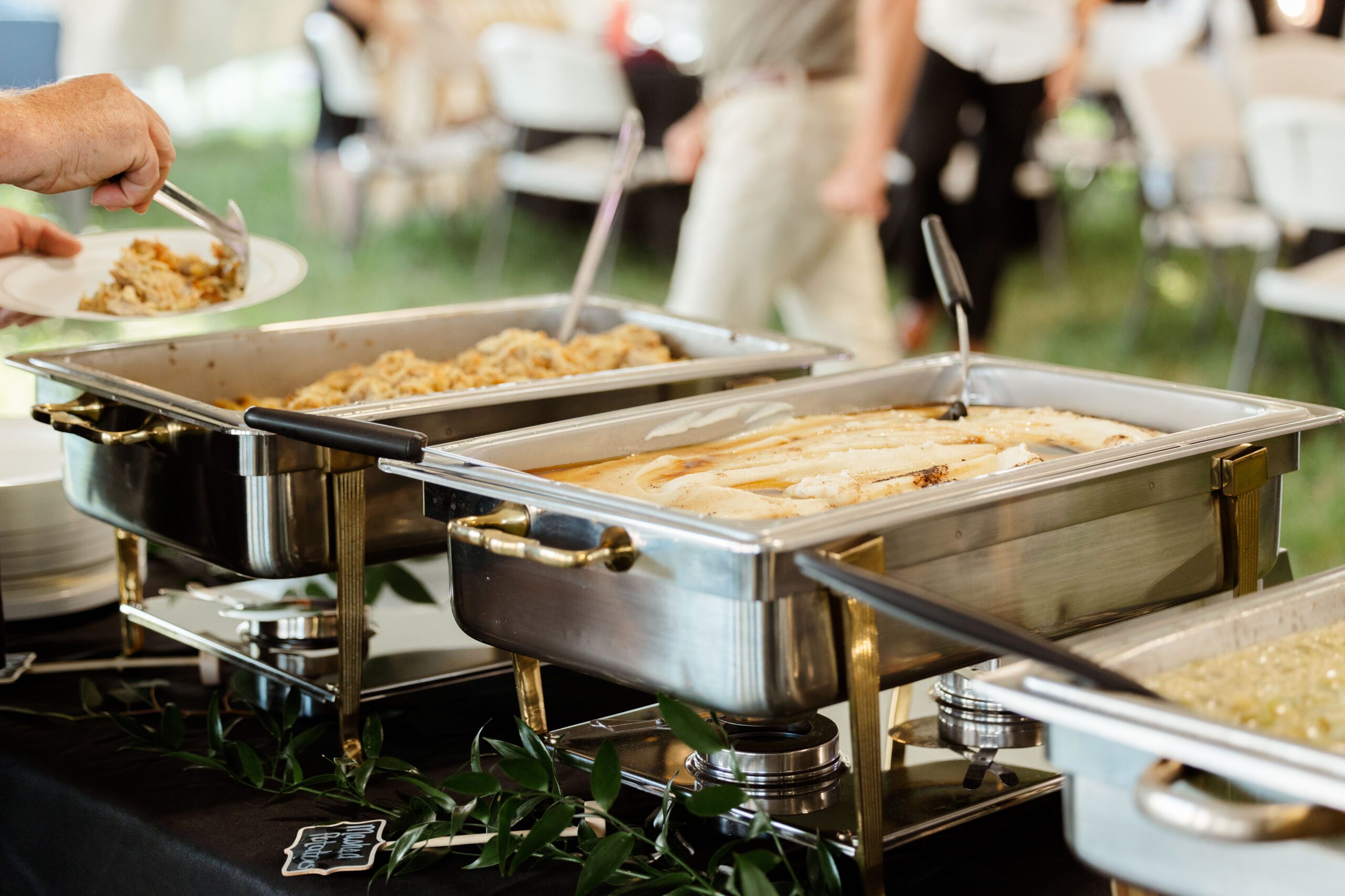 Image of a buffet table featuring silver chafing dishes containing various dishes. A person is serving food onto a plate, while additional plates and seating are visible in the background, indicating a catered event or gathering.