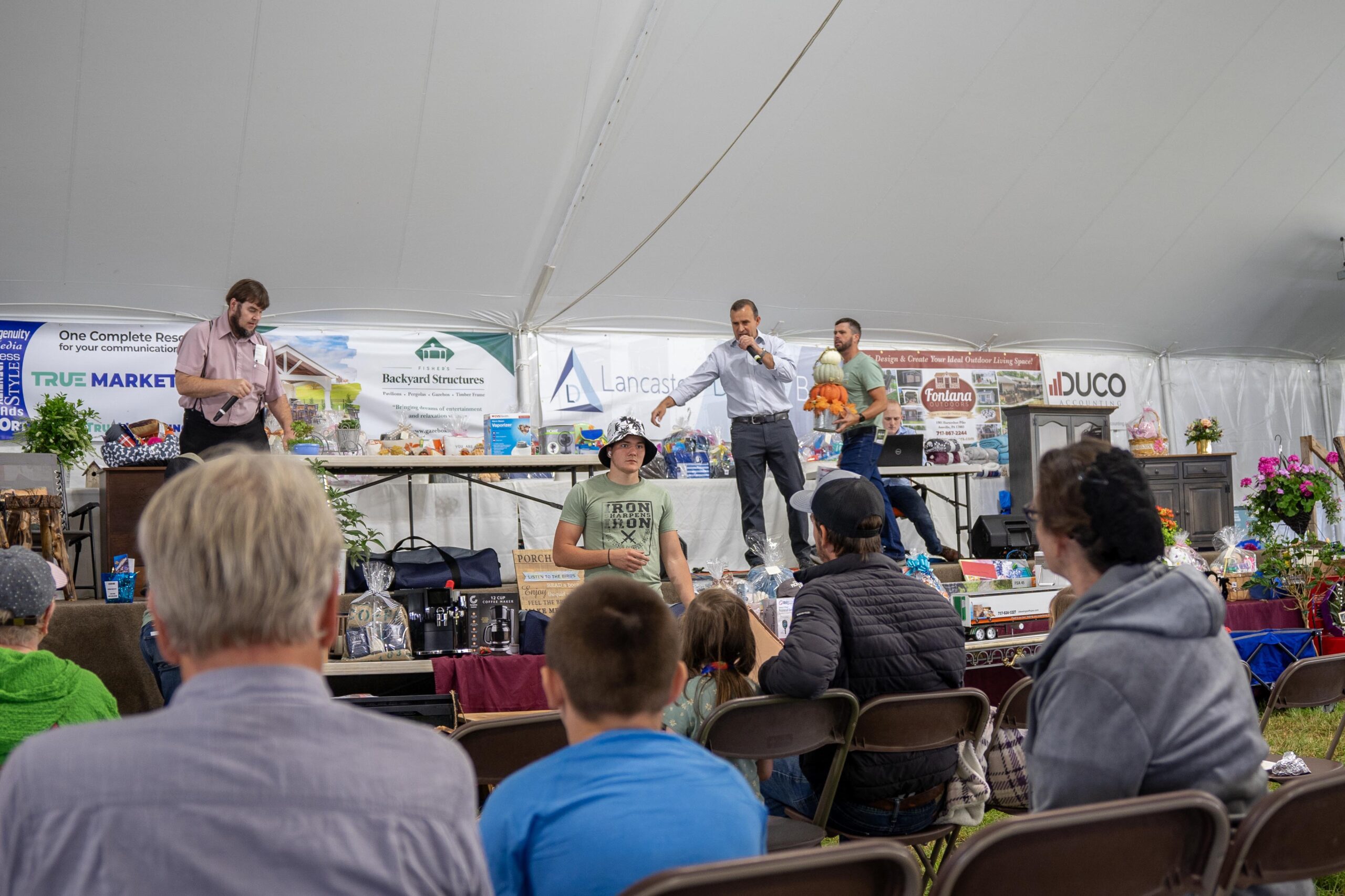 Audience attending a live auction event under a tent, featuring a presenter on stage discussing items for bidding while participants observe. The display includes various items for auction, surrounded by greenery and colorful decorations.