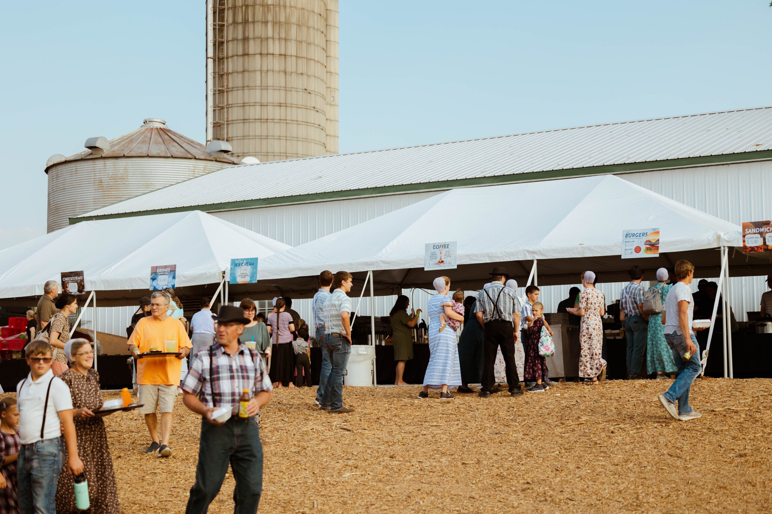 Crowd enjoying food stalls at a rural festival, with several tents offering burgers, coffee, and ice cream, set against a barn and silo backdrop.