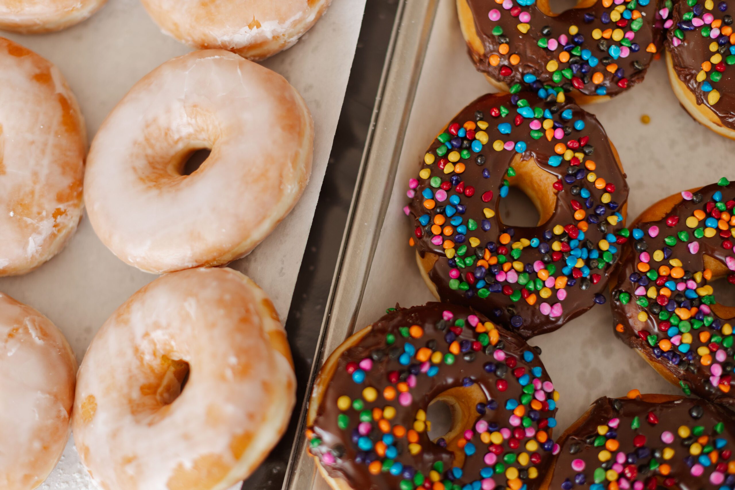 Assorted donuts featuring classic glazed rings and colorful chocolate-covered varieties topped with sprinkles, displayed on a baking tray.