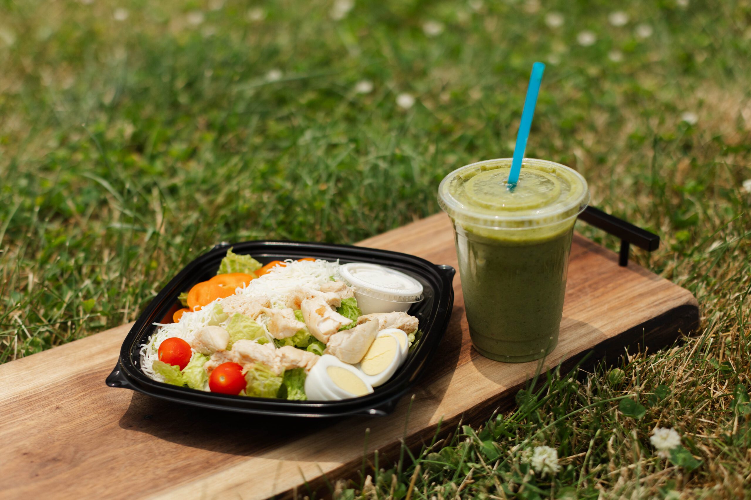 Healthy salad bowl with grilled chicken, mixed greens, cherry tomatoes, and sliced boiled eggs, accompanied by a green smoothie on a wooden board outdoors.