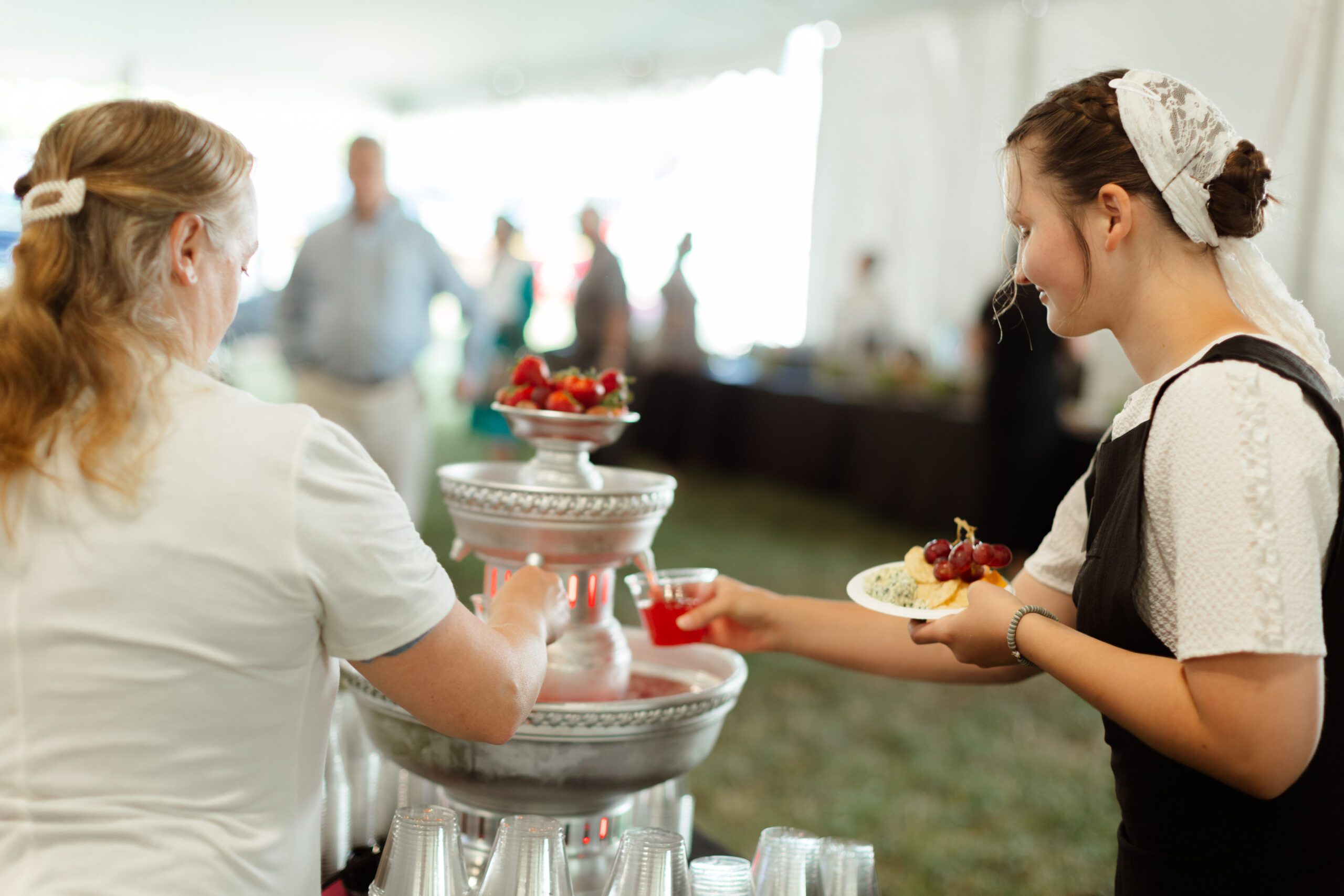 A woman serves a refreshment from a drink fountain at an outdoor event, while another guest holds a plate with fresh fruit and snacks. The setting features a tent filled with people enjoying the gathering.