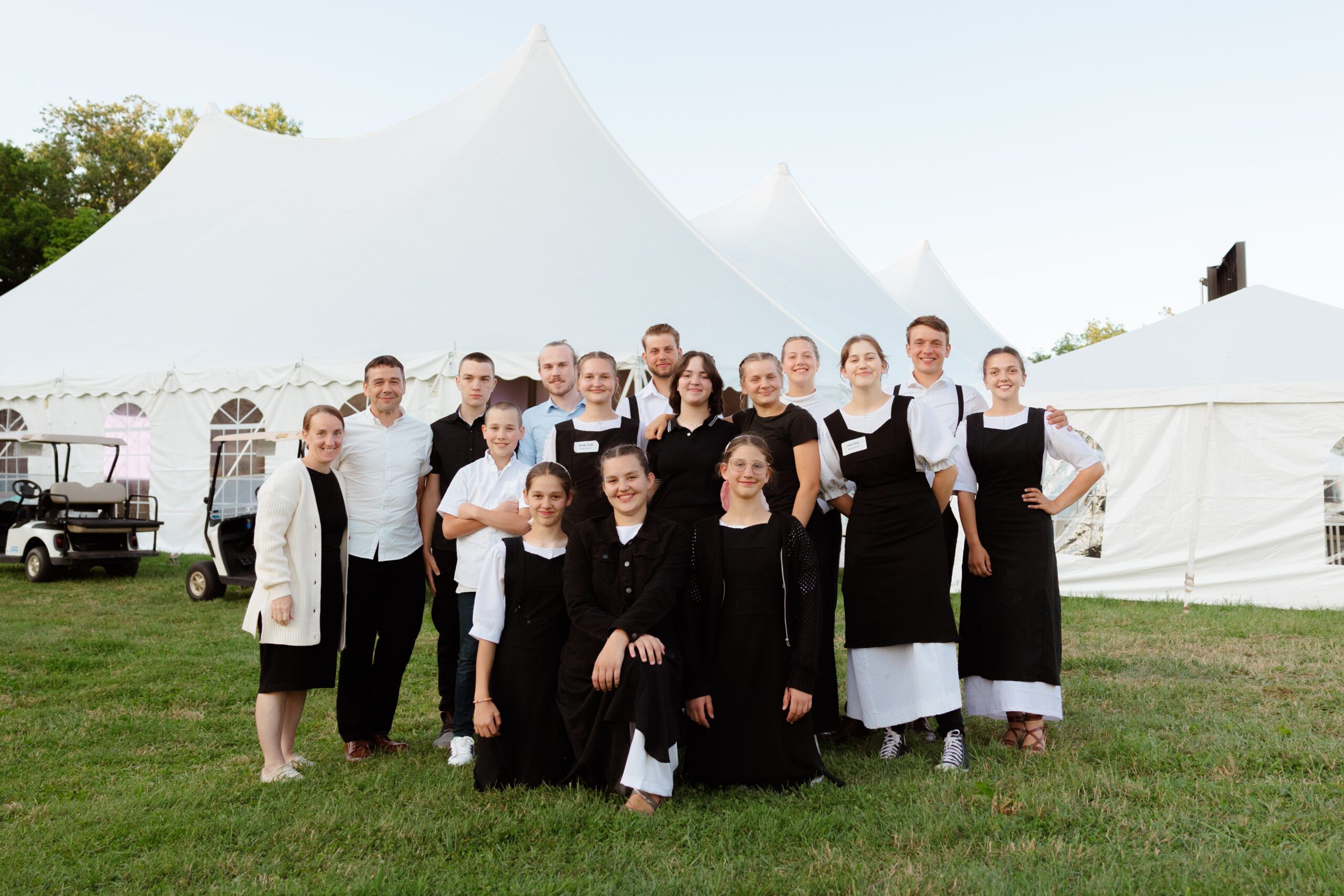 Group photo of a diverse team of event staff wearing black and white uniforms, smiling together in front of a large white tent at an outdoor venue, showcasing teamwork and hospitality.
