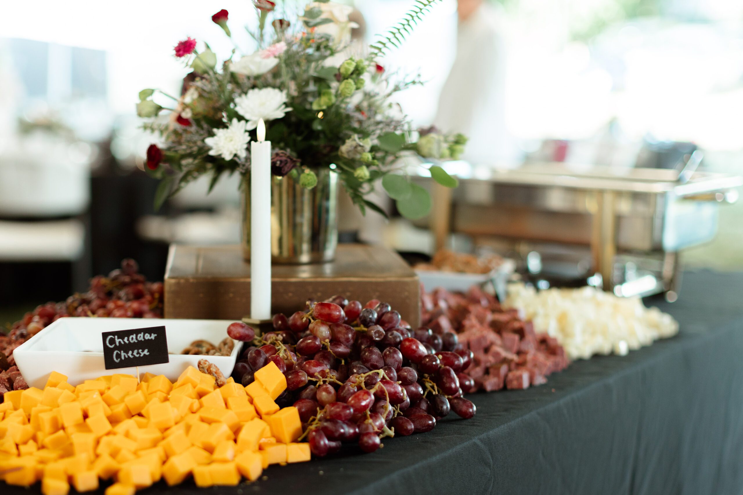 Cheese and charcuterie table featuring blocks of cheddar cheese, fresh grapes, and assorted meats, adorned with a floral centerpiece and a candle, creating an elegant ambiance for events and gatherings.
