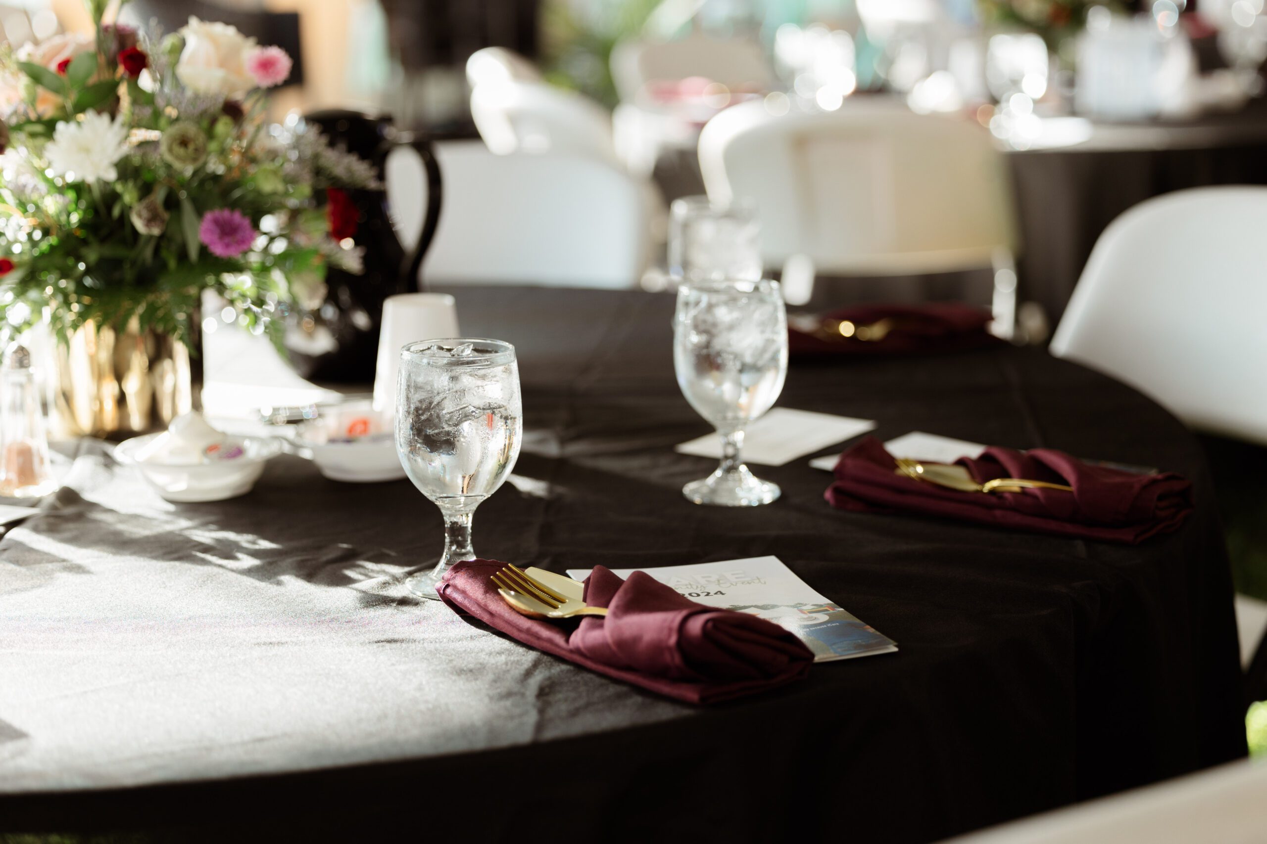 Elegant dining setup featuring water glasses, gold cutlery, and burgundy napkins on a black tablecloth, with a vibrant floral centerpiece, suitable for weddings or formal events.