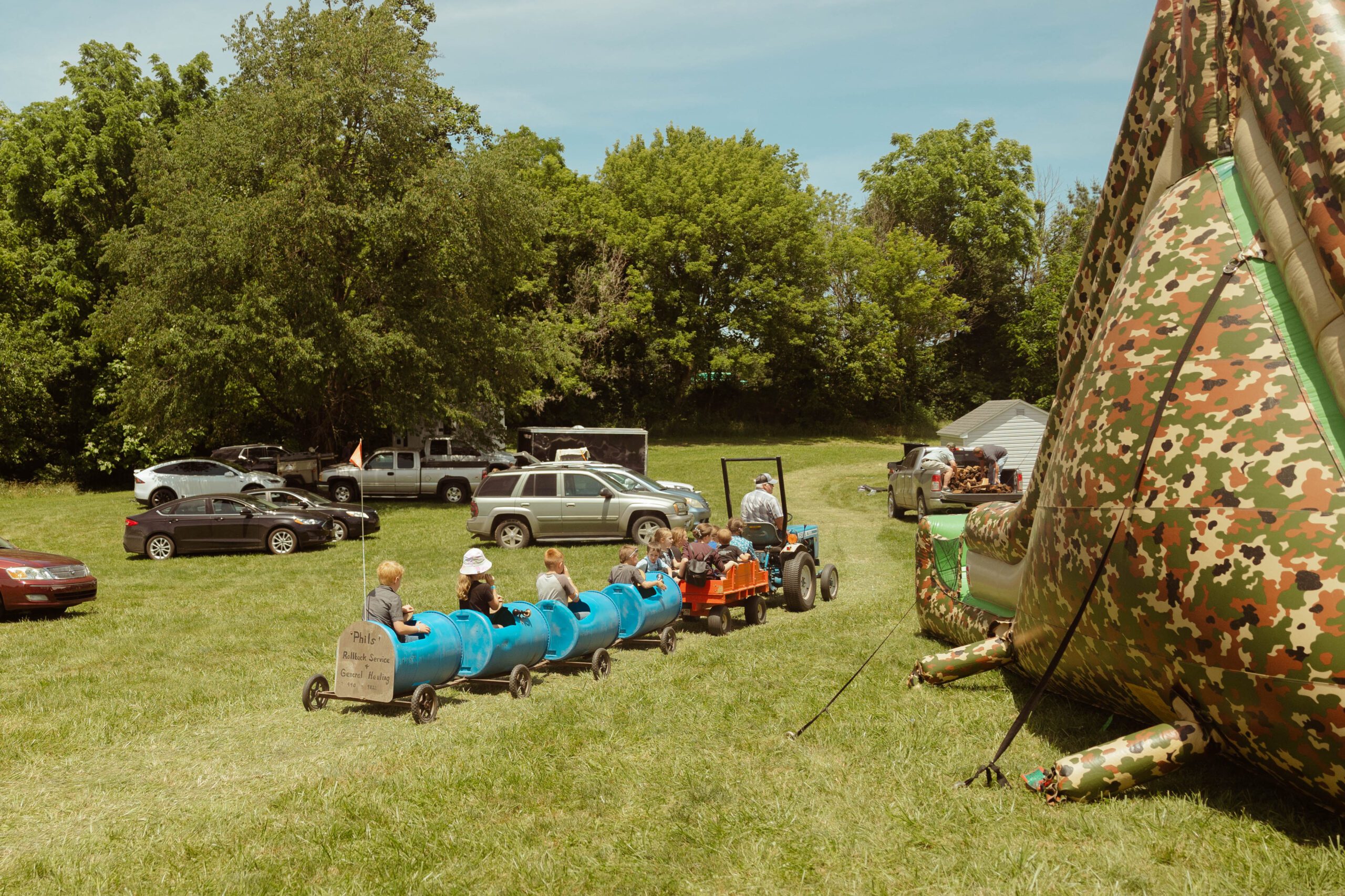 Children enjoying a tractor-pulled ride in blue barrels at an outdoor event, with parked cars and a large inflatable structure in the background, surrounded by lush greenery.
