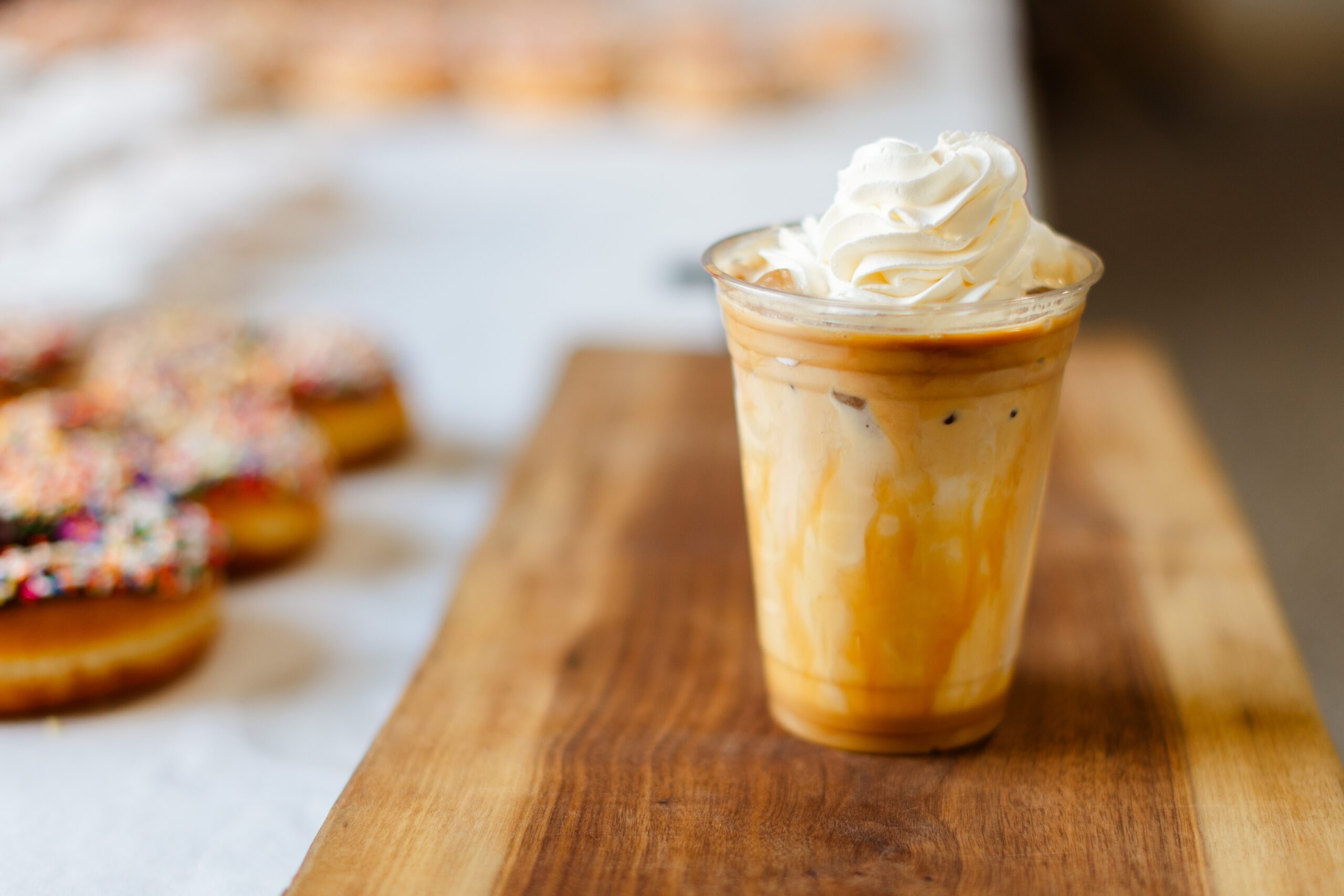 Iced coffee topped with whipped cream, served in a clear cup on a wooden surface, with colorful frosted donuts in the background.