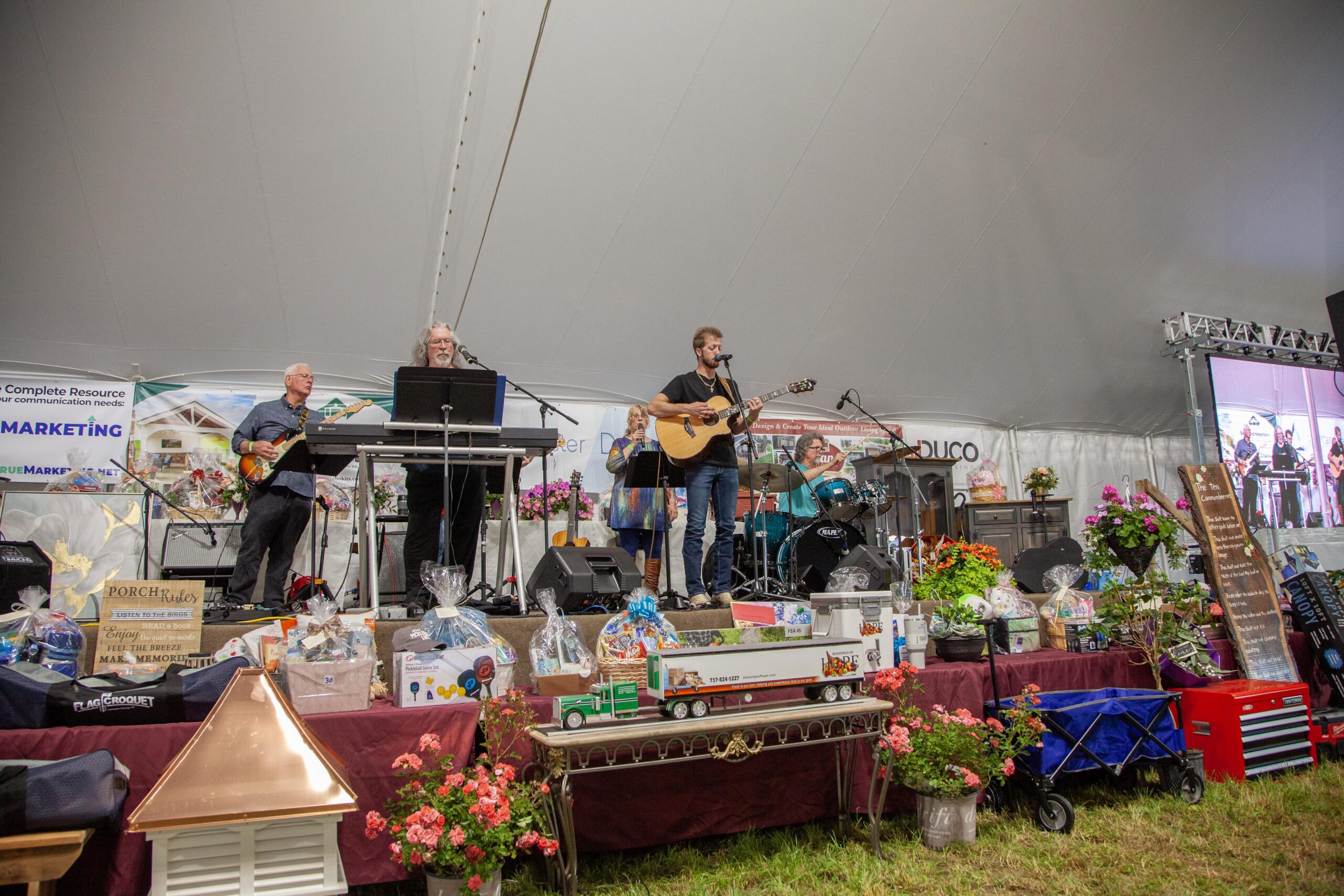 Live band performing on stage at a festival, featuring musicians playing guitar, keyboard, and drums. The backdrop displays promotional banners, while tables in the foreground showcase items for a raffle or auction, surrounded by floral decorations.