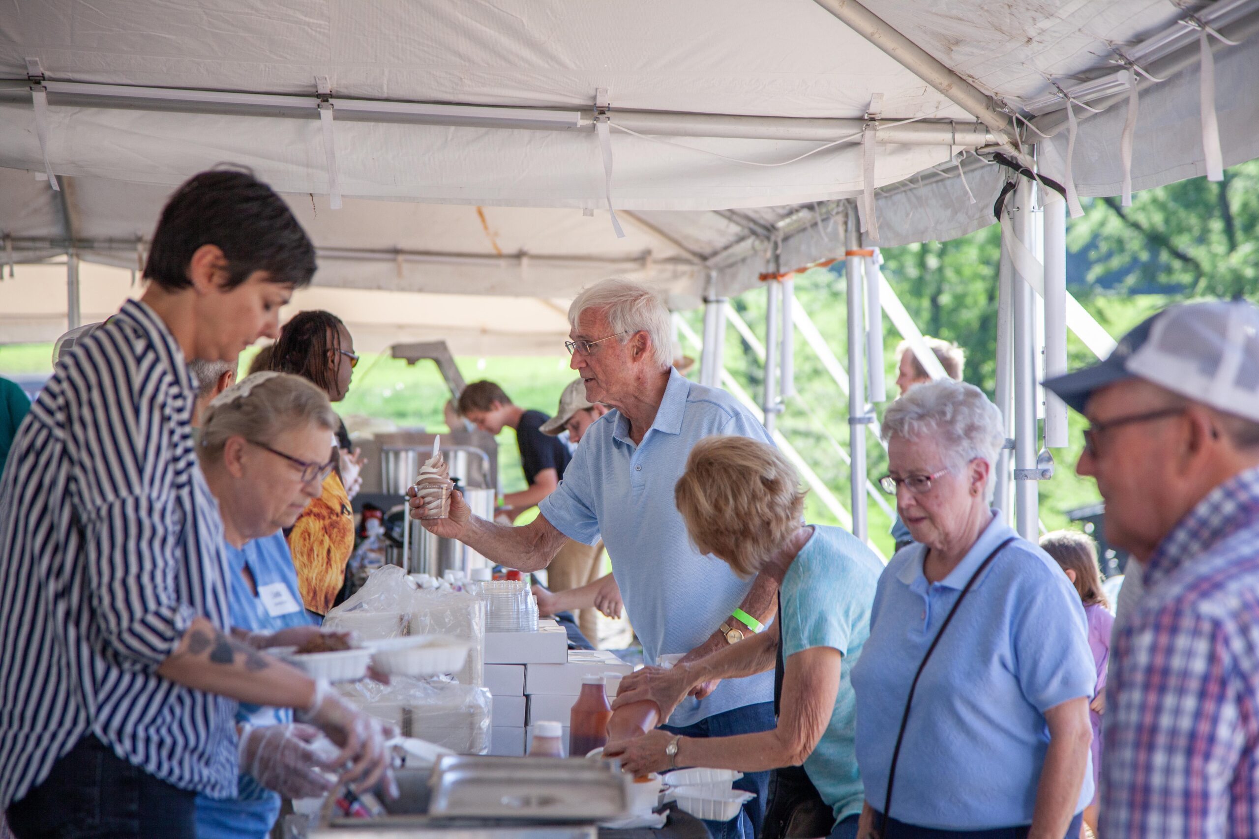 A lively outdoor community event where volunteers of various ages serve food from a buffet setup under a large tent. People are engaged in conversation while receiving meals, showcasing a sense of camaraderie and community spirit.