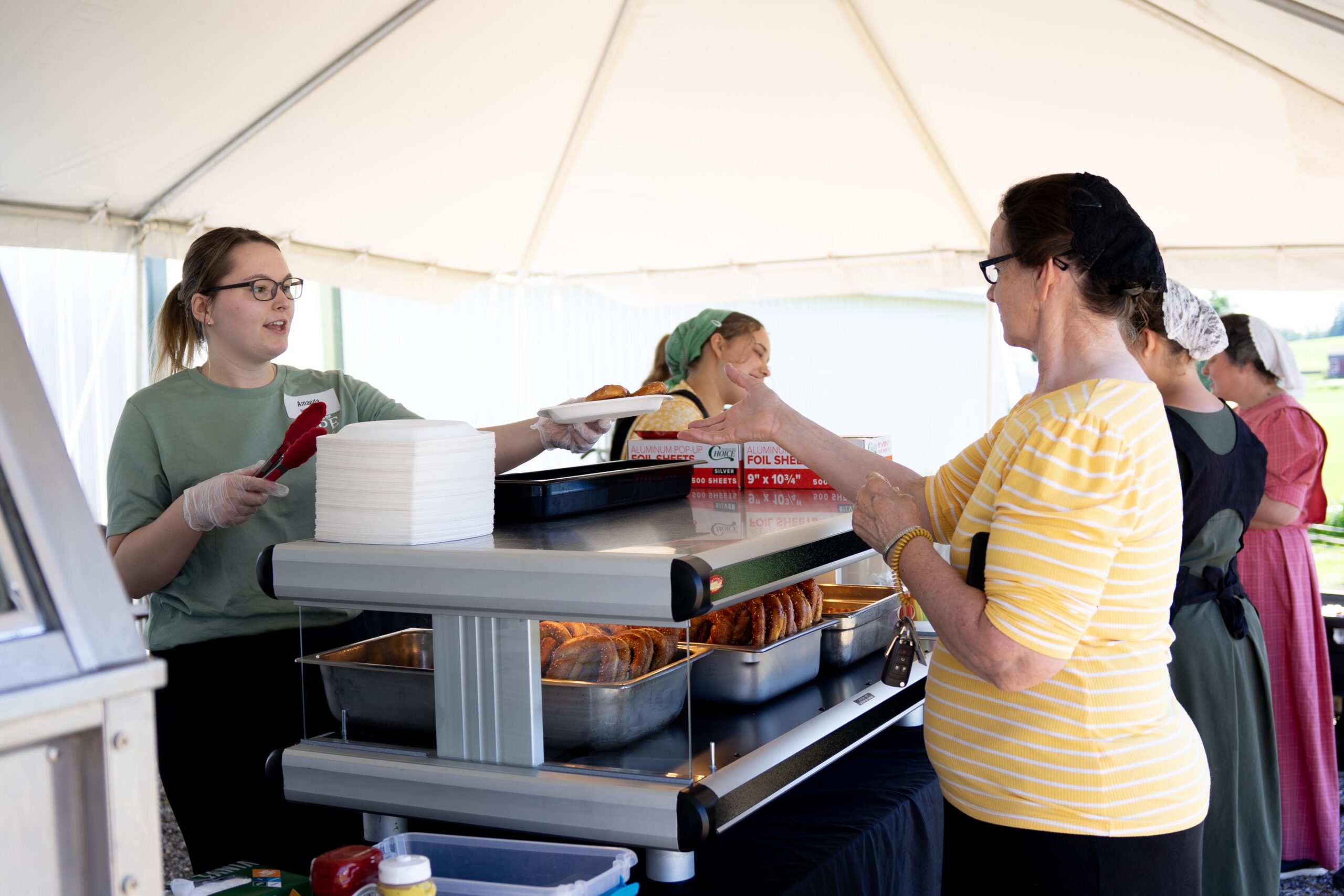 Staff serving food at a bustling outdoor event under a tent, with a woman in a striped shirt receiving her meal. The setup includes metal food trays, plates, and utensils, highlighting a community gathering focused on traditional cuisine.