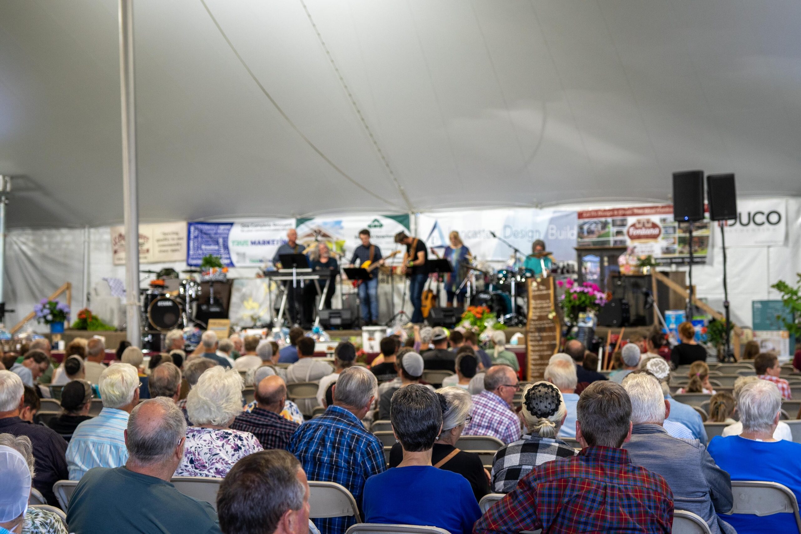 Audience enjoying a live musical performance under a large tent, featuring a diverse band playing various instruments. Colorful floral arrangements enhance the stage area, while banners from local businesses are displayed in the background.