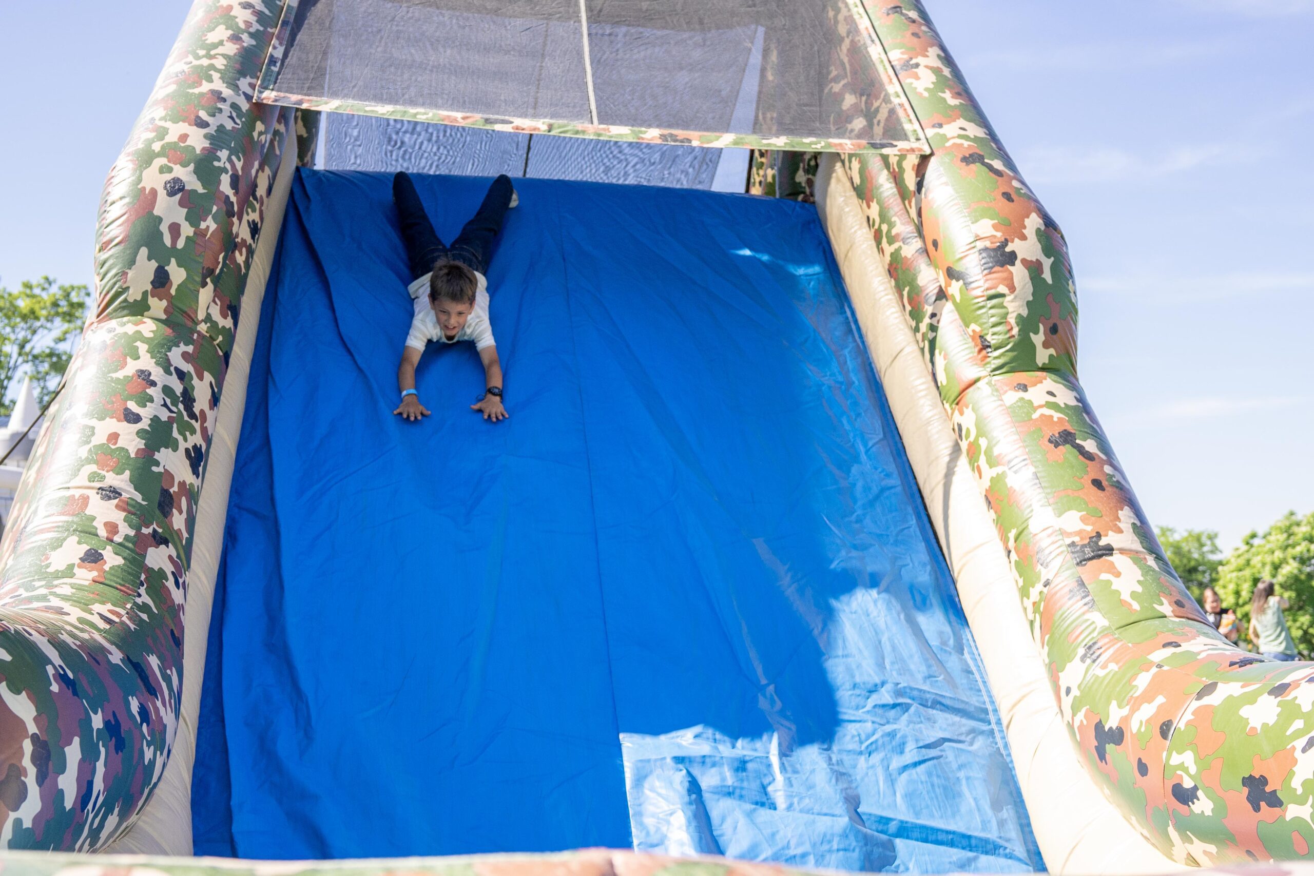 Child sliding down a colorful inflatable slide at an outdoor event, with a bright blue surface and a camouflaged border. Trees and a clear sky visible in the background.