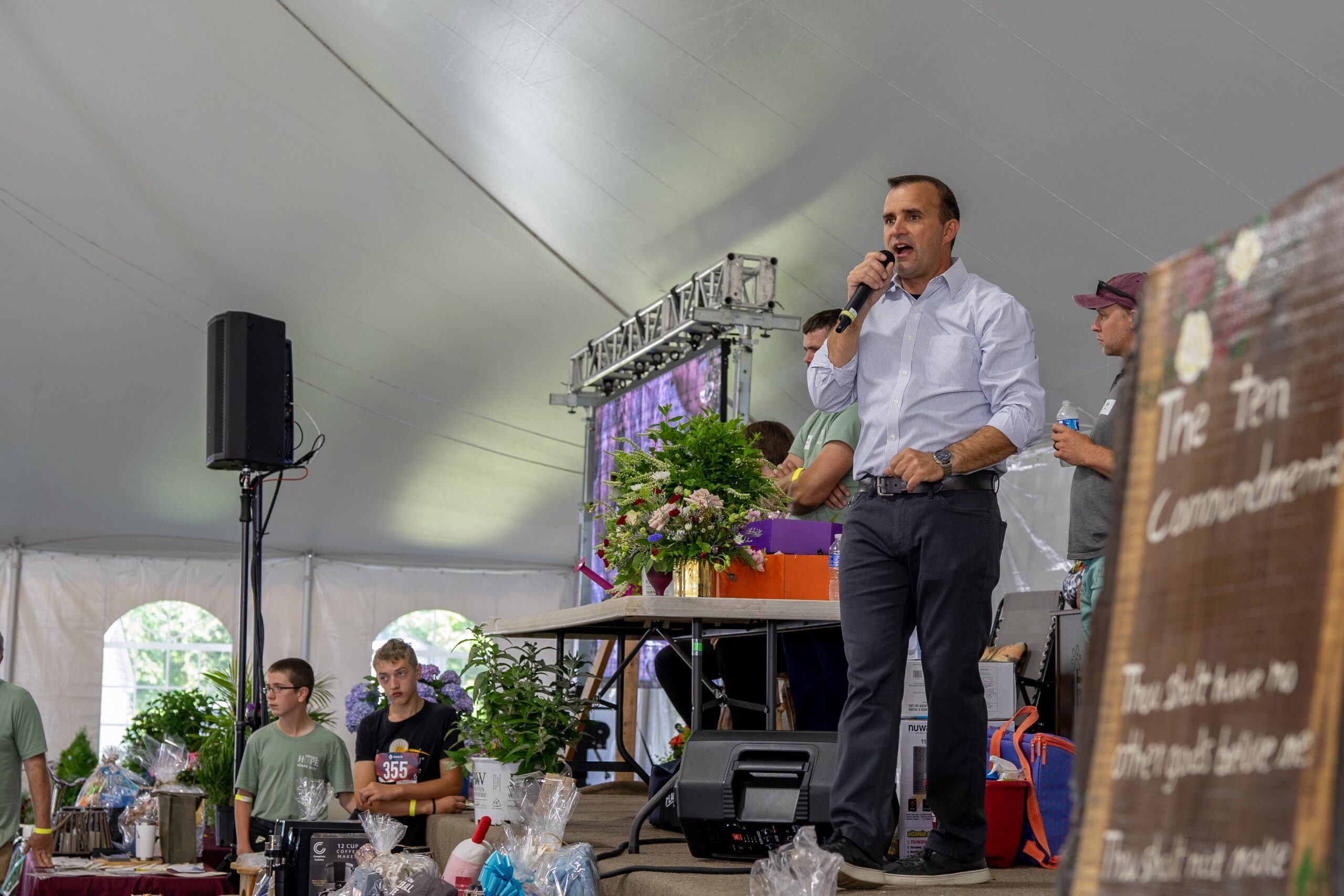 Speaker addressing an audience at a community event under a large tent, with a microphone in hand and surrounded by plants and gift baskets on display. Participants, including teenagers, appear engaged in the background.