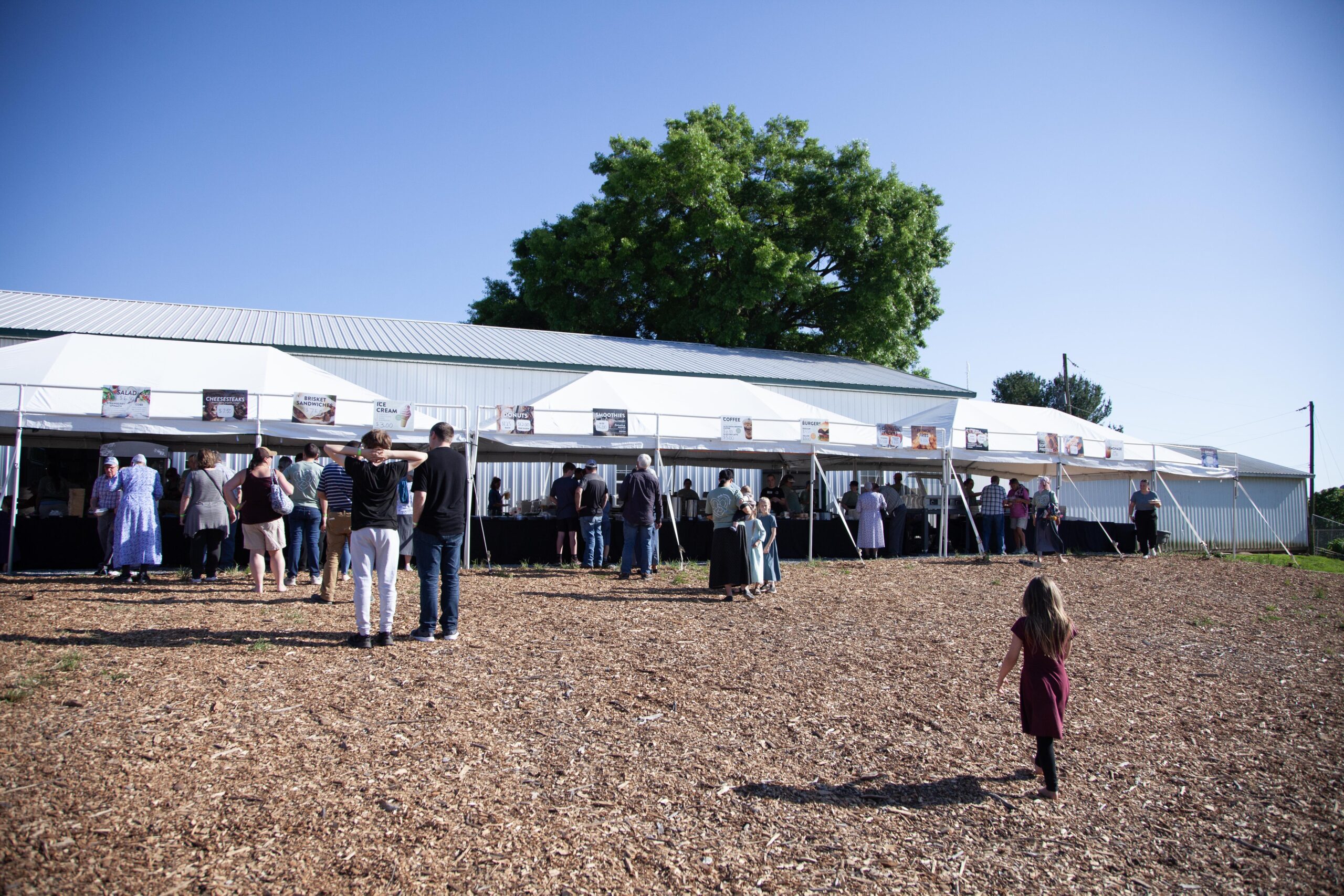 Crowd of people attending an outdoor food event near a barn, with several vendor tents displaying menu items like ice cream and baked goods under a clear blue sky. A young girl walks through the wood-chip covered ground.