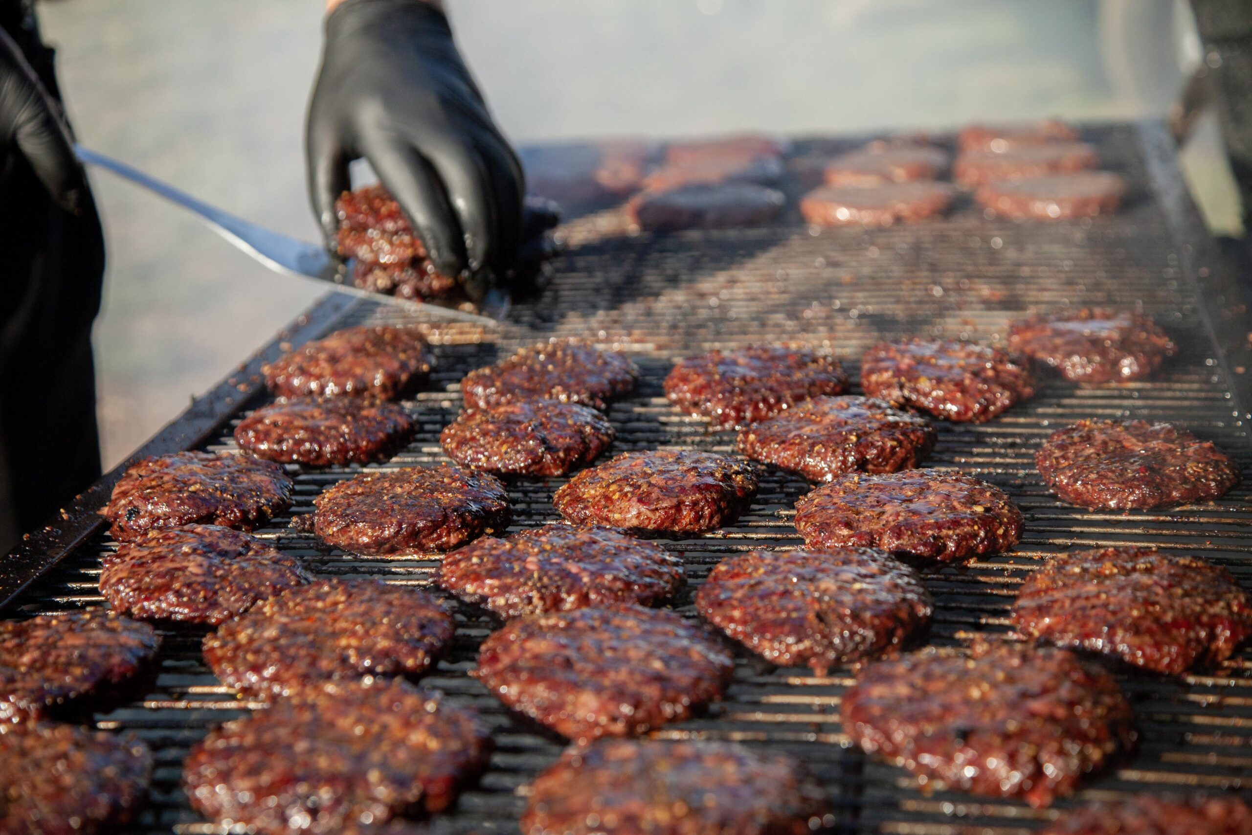 Close-up of hamburgers sizzling on a grill, with smoke rising and a hand in black gloves using a spatula to flip a patty, showcasing a summer BBQ scene.
