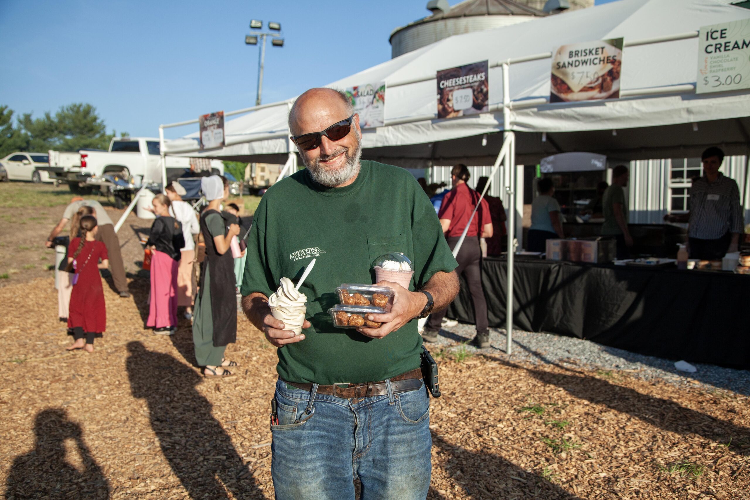 A smiling man holding a soft-serve ice cream cone and a container of baked goods stands in front of a food stall at an outdoor market. Behind him, a variety of food items such as cheesesteaks and brisket sandwiches are displayed. The scene is lively with people in casual attire and a sunny sky.
