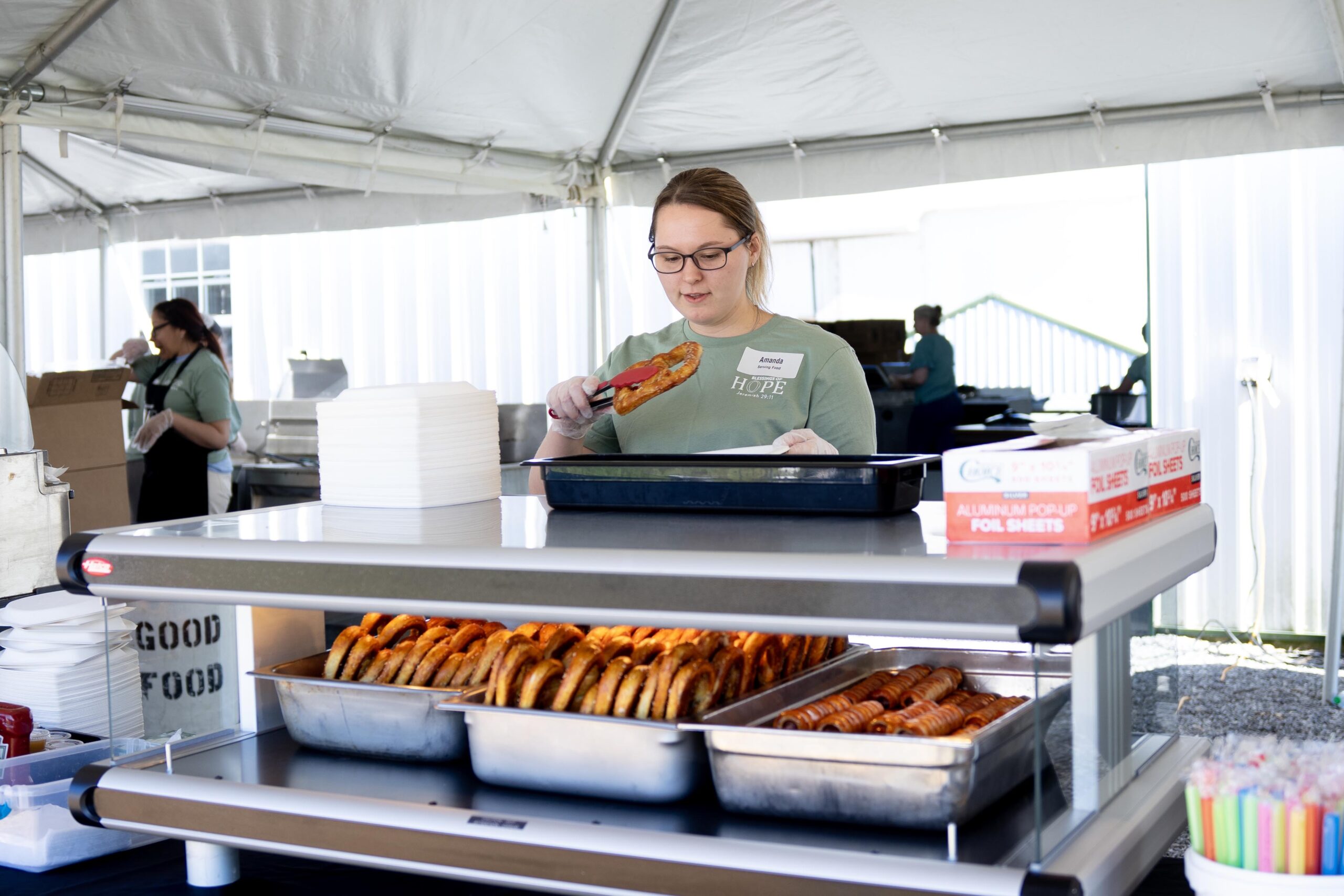 Woman serving food at outdoor event booth with pretzels and paper plates on display.