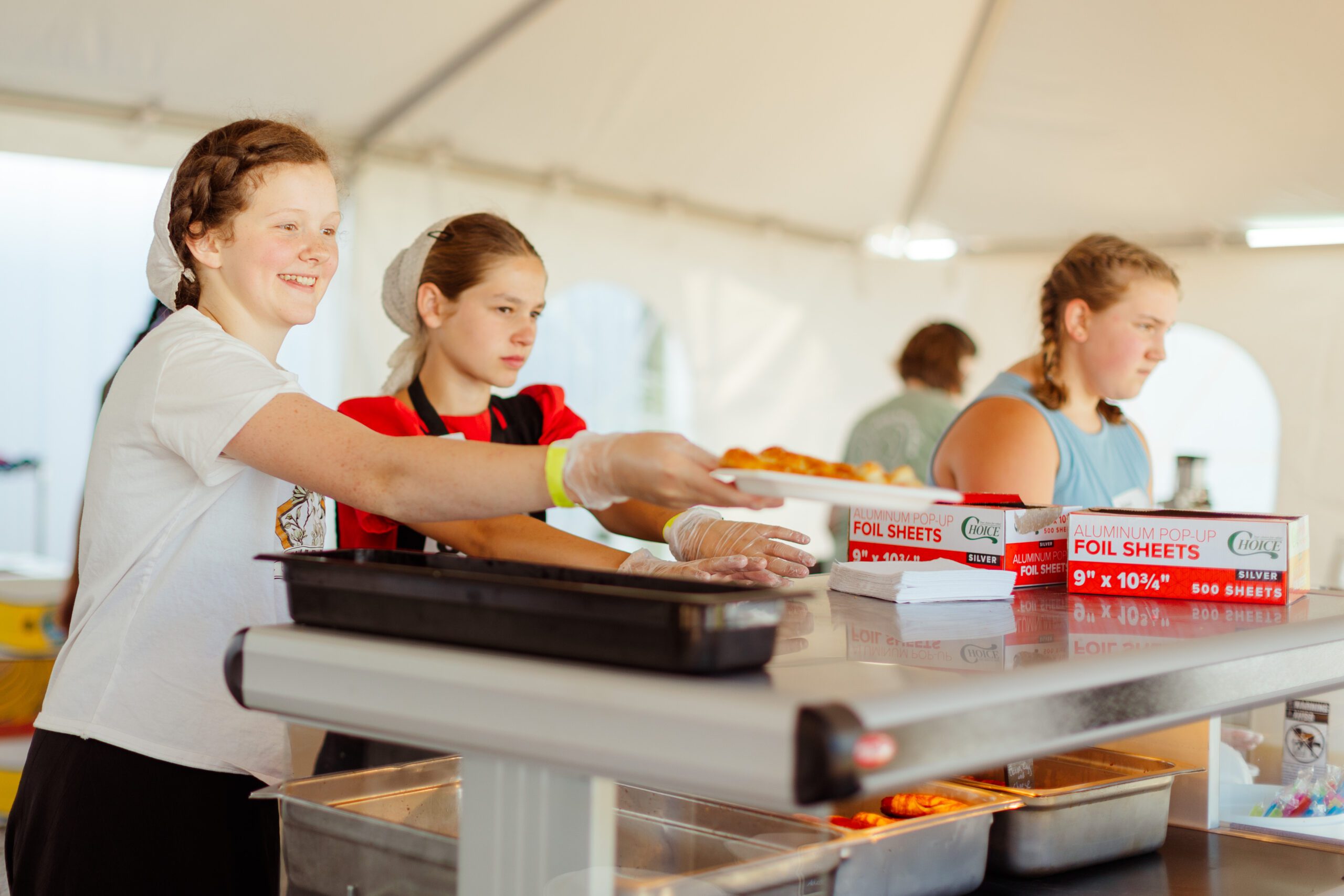Volunteers serving food at a community event under a tent, with one girl smiling as she hands out hot dishes, while two others assist in the background.
