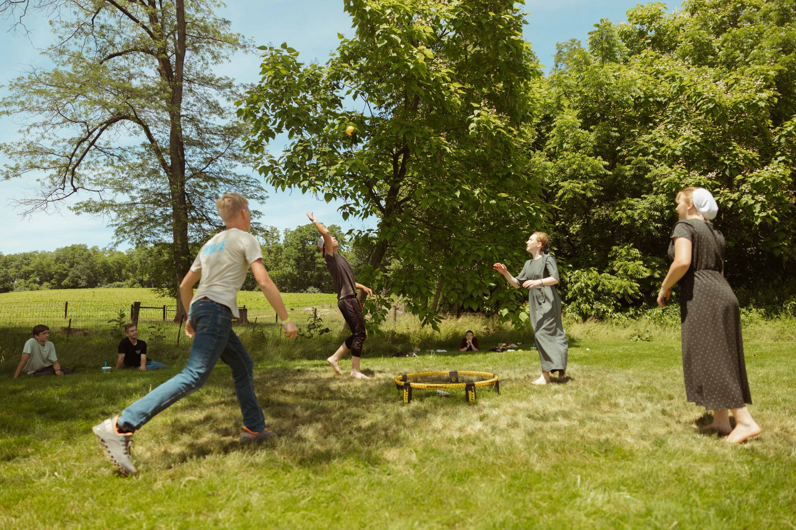 A group of young people playing a lively game of spikeball outdoors, surrounded by lush greenery and a clear blue sky. Some participants are jumping and reaching for the ball, while others watch from a grassy area. The scene captures the joy of outdoor summer activities.