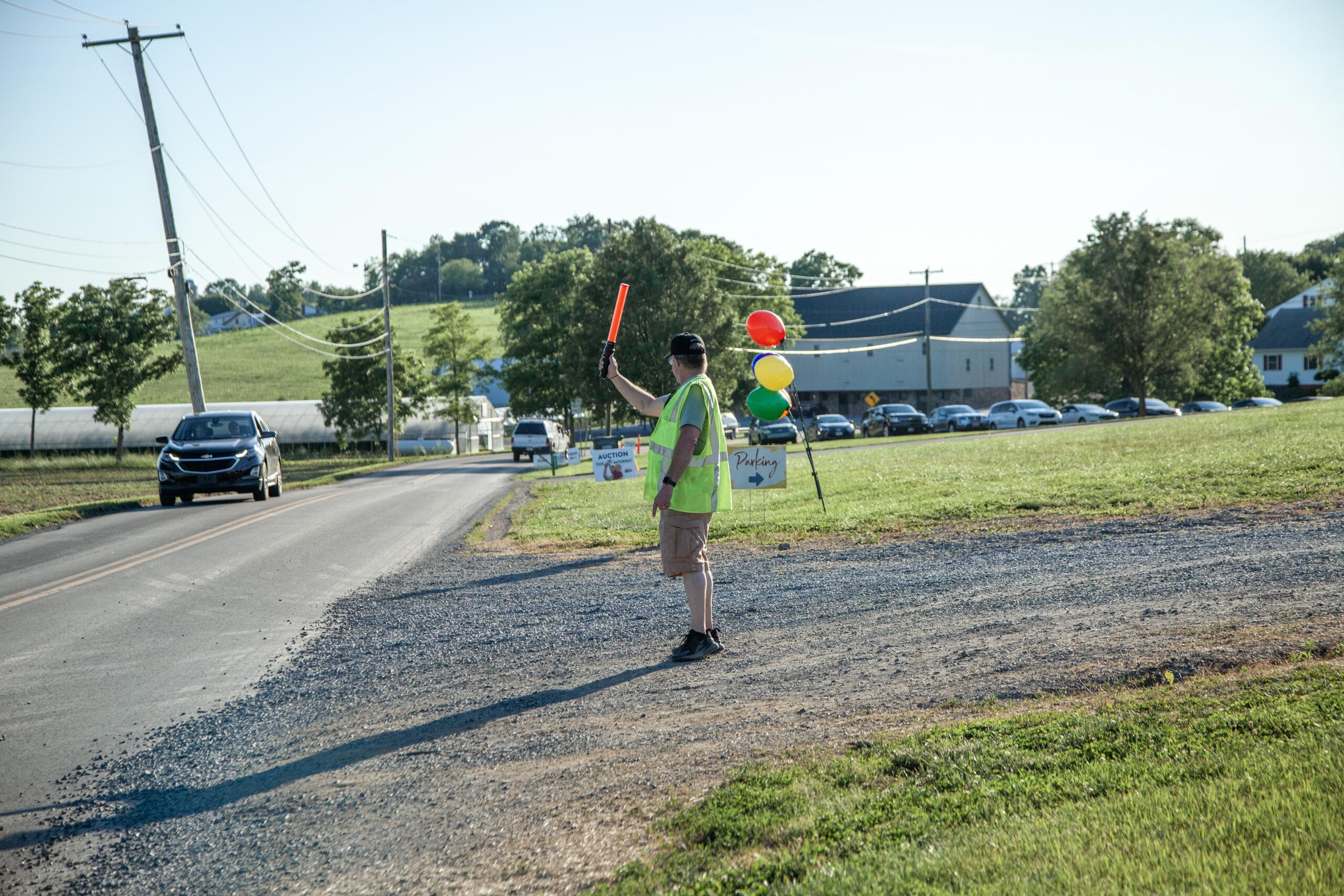 Traffic coordinator directing vehicles with a signaling wand at an outdoor event, featuring colorful balloons and signs for parking and an auction in the background.