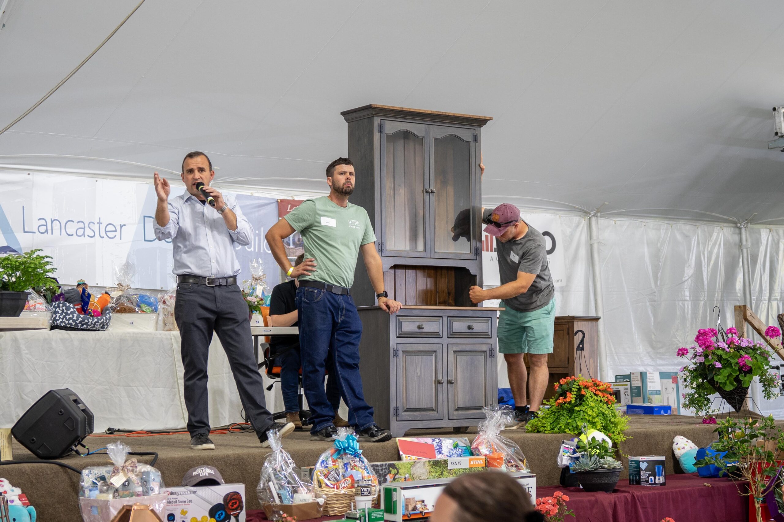 Men engaged in a furniture auction at a community event, featuring a wooden cabinet on stage, with decorations and items for sale in the foreground.