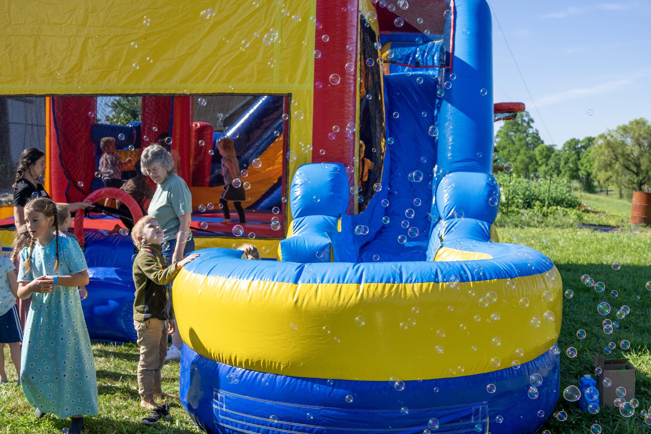 Children playing near a colorful inflatable bounce house with bubbles in the air, set in a green outdoor environment. Two kids are interacting with the bounce house, while adults supervise in the background.