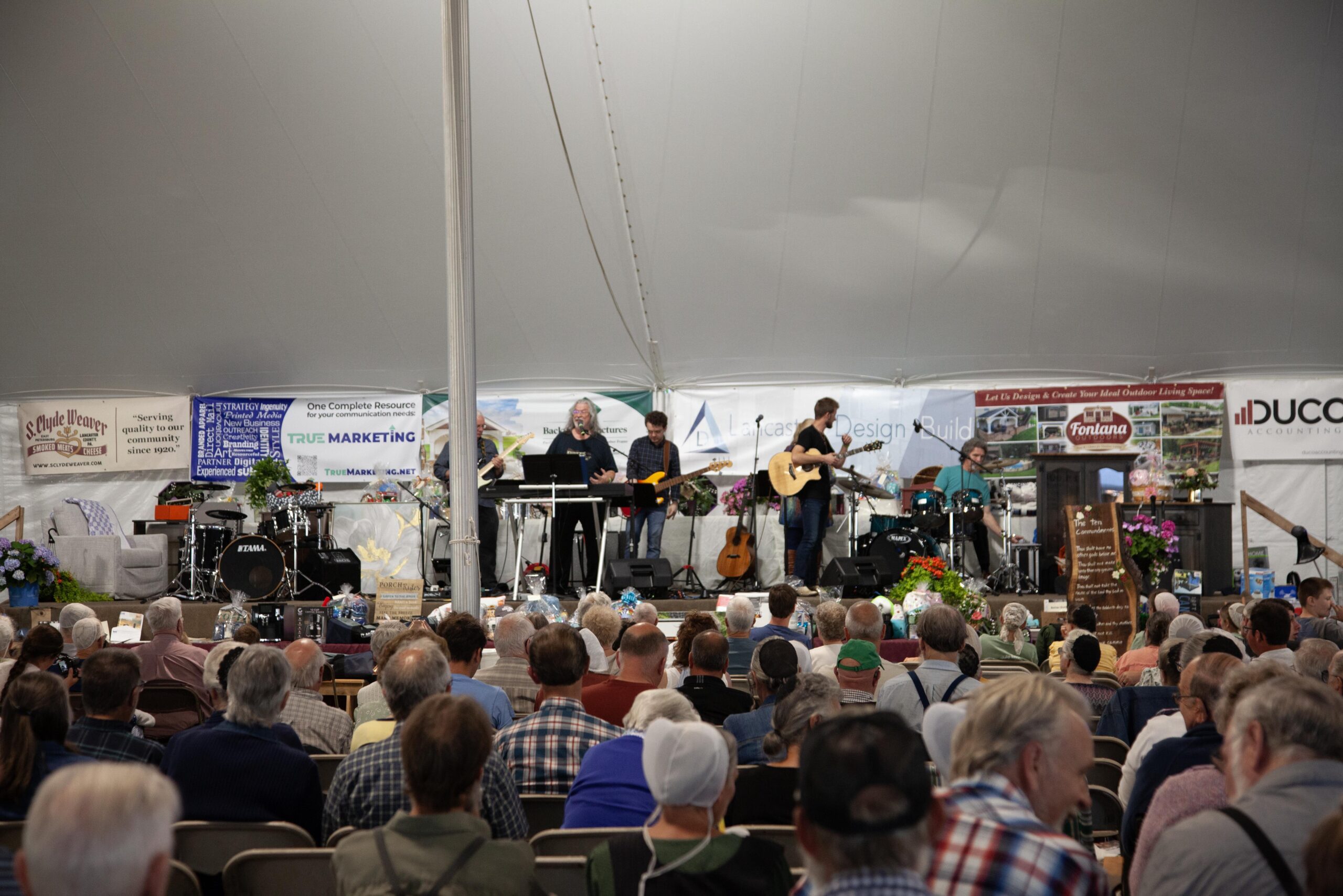 Live musical performance in a large tent featuring a band with various instruments, including guitars and drums, entertaining a seated audience. Event banners displaying local businesses and community support are visible in the background, creating a vibrant atmosphere.