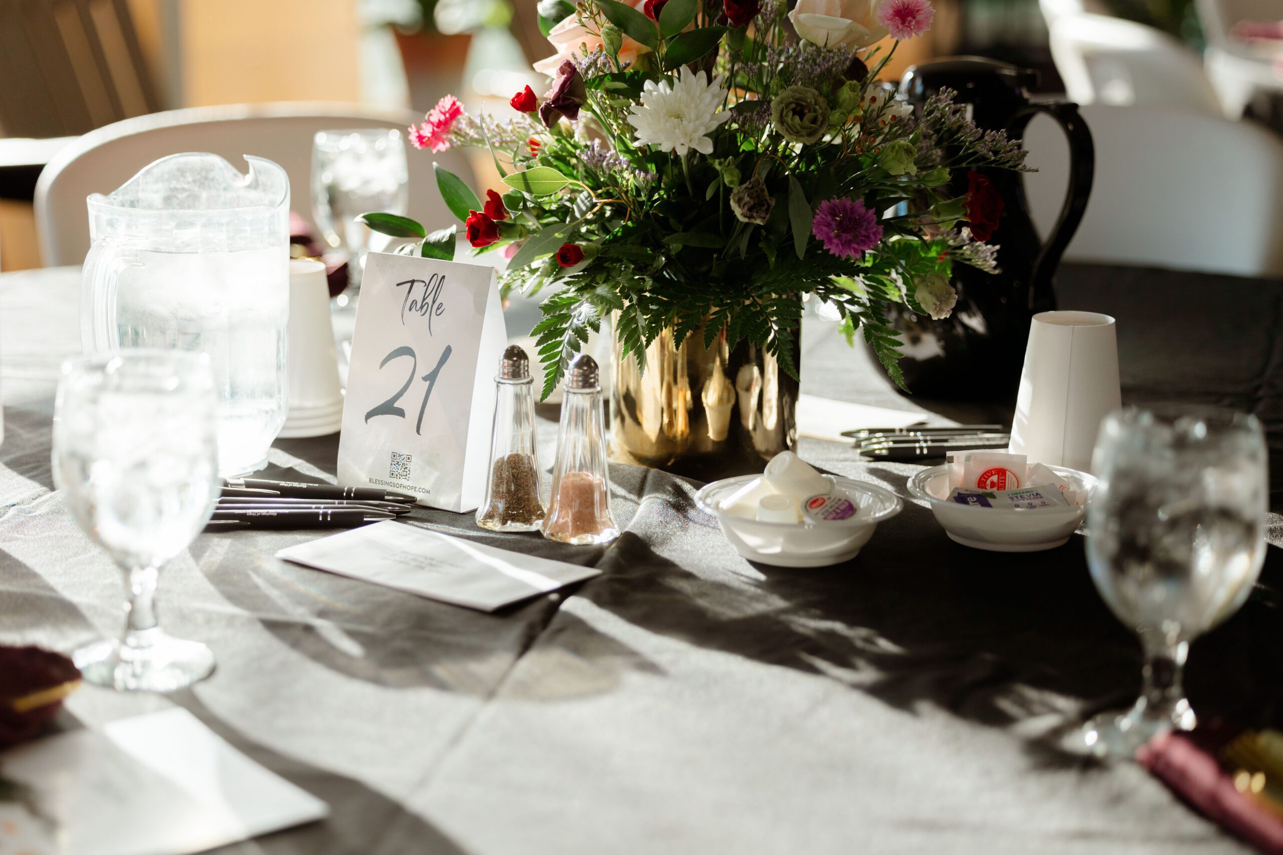 Table setting for an event featuring a decorative floral centerpiece, a table number sign labeled "Table 21," glassware, salt and pepper shakers, and condiments on a black tablecloth.