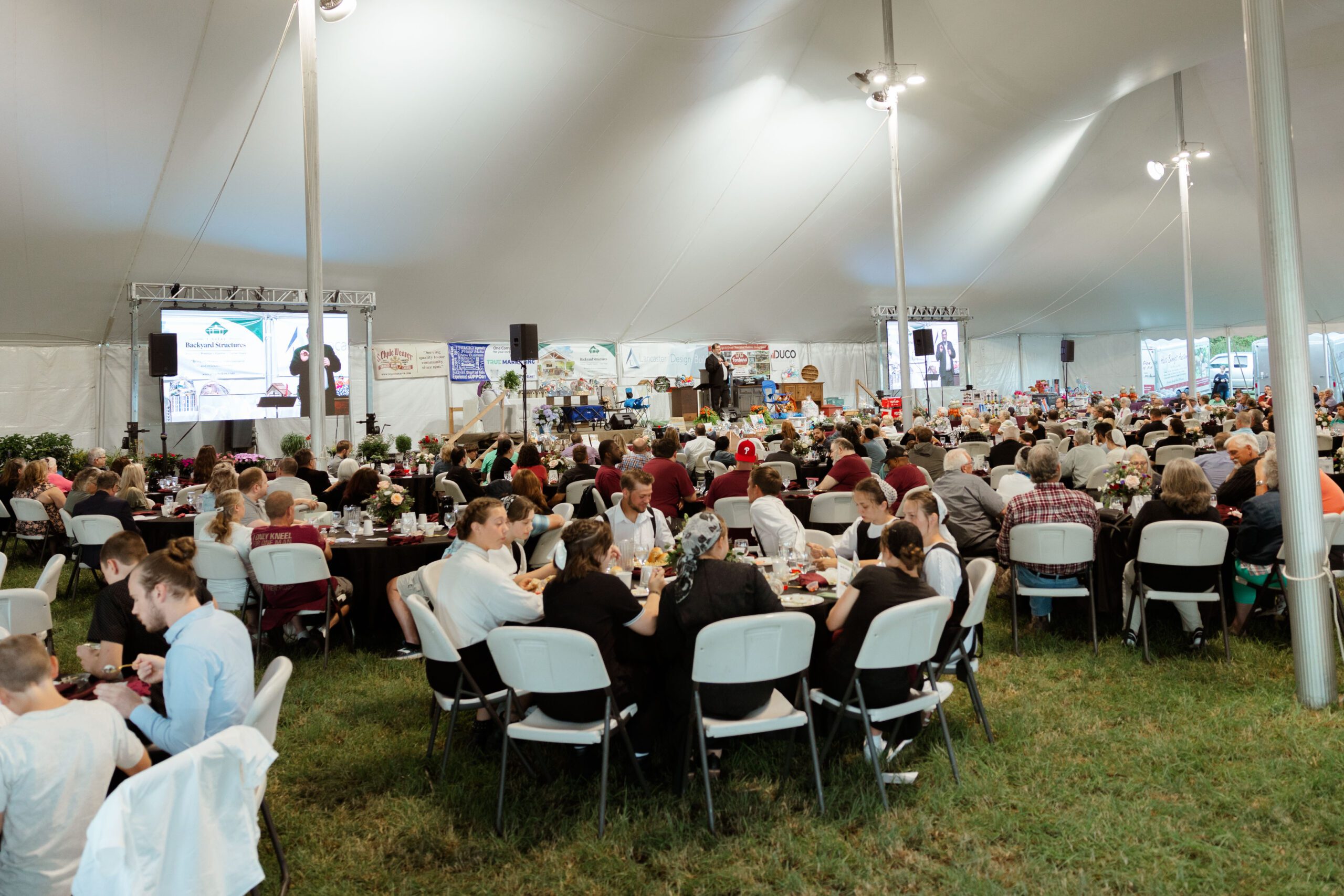 Large gathering under a tent featuring attendees seated at tables enjoying a meal, with a speaker on stage and various event banners in the background.