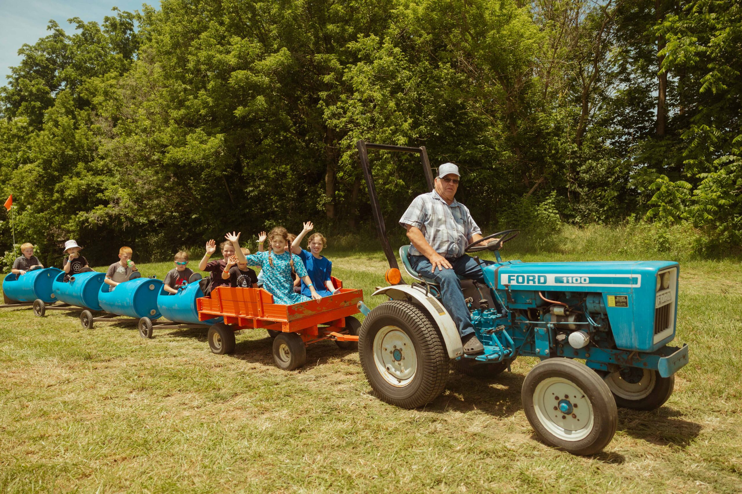 A man driving a blue Ford tractor pulls a colorful cart filled with children waving and enjoying a fun outdoor activity amidst lush greenery on a sunny day.