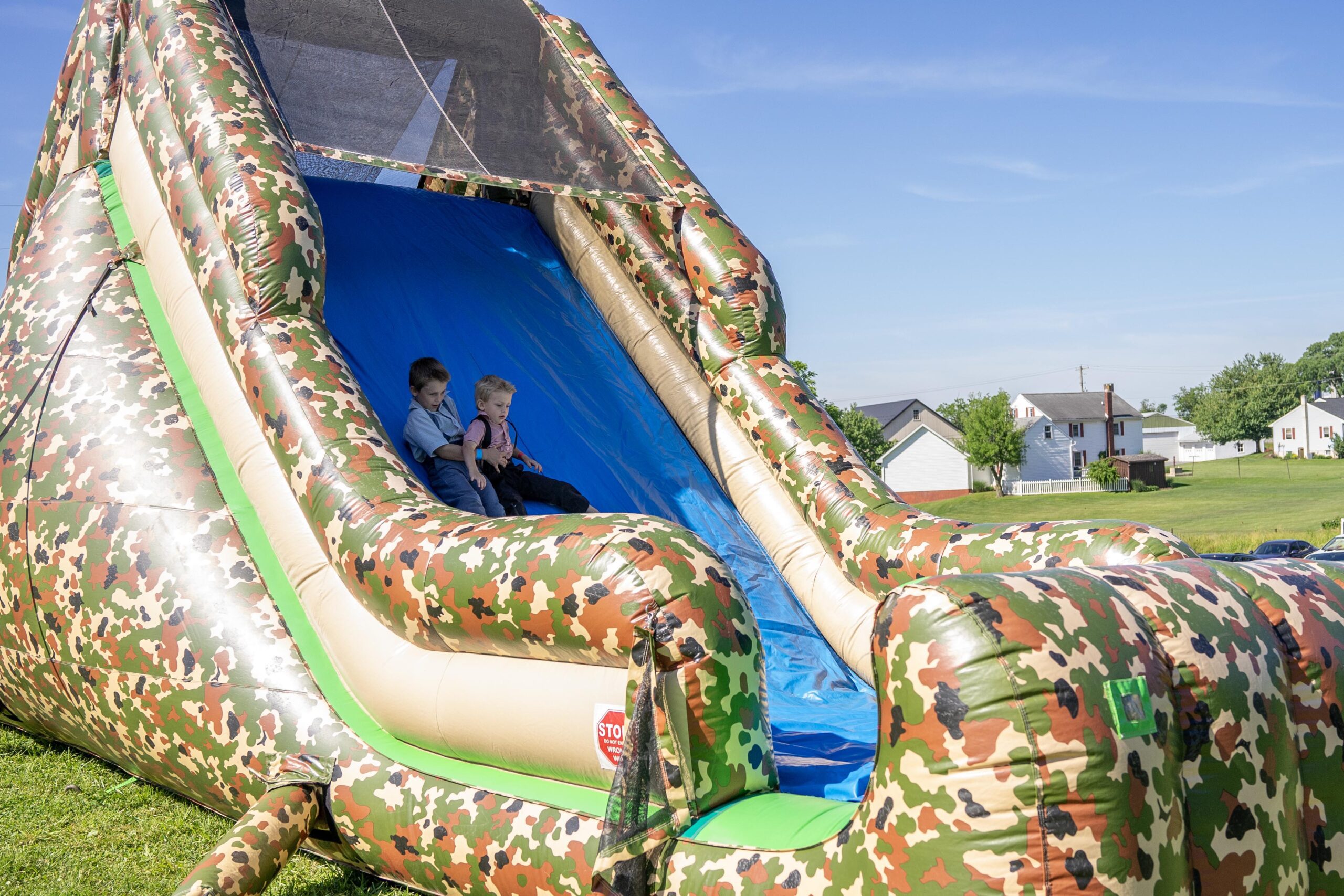 Kids sitting at the top of a colorful inflatable camouflage slide, ready to enjoy a fun day outdoors in a park setting.