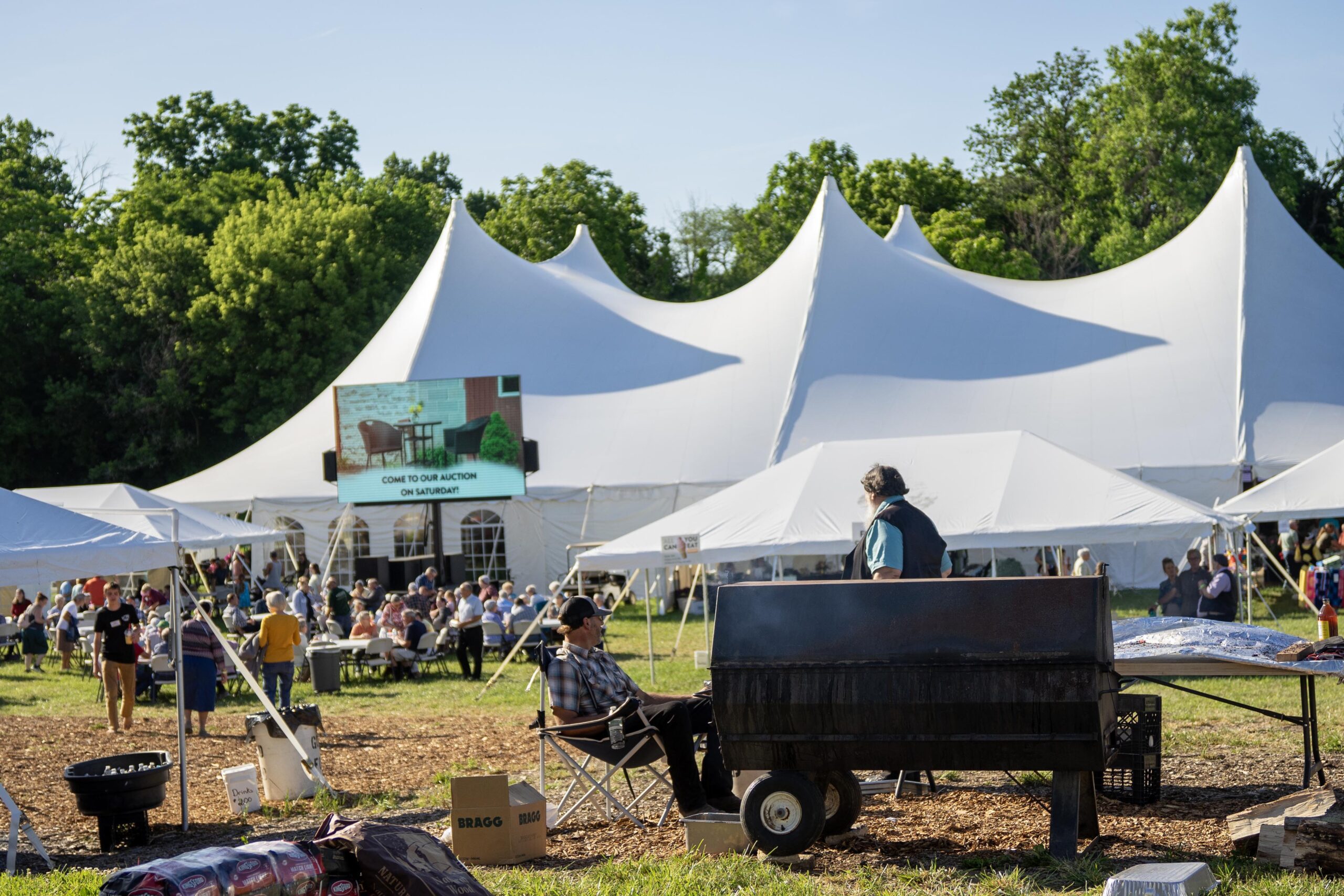 Crowd enjoying an outdoor event with a large tent, featuring an auction announcement on a screen, and a barbecue setup in the foreground.