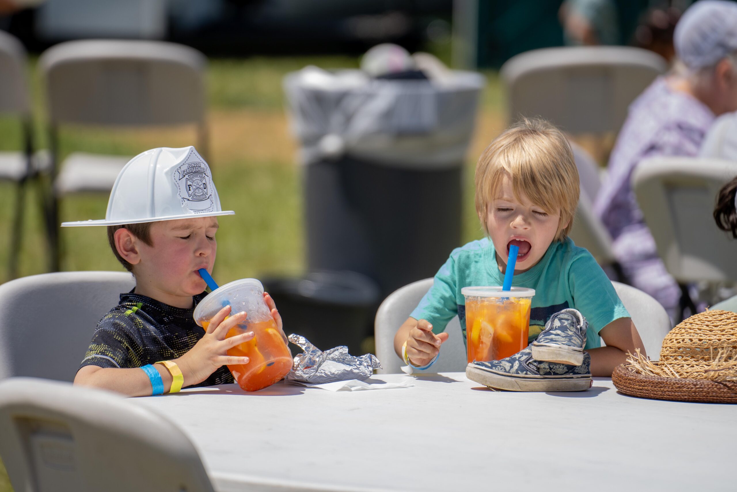 Two young boys sitting at a table, enjoying colorful drinks with large straws, on a sunny day. One boy is wearing a fireman’s helmet, while the other sports a turquoise shirt. They are surrounded by outdoor seating and foliage, highlighting a festive atmosphere.