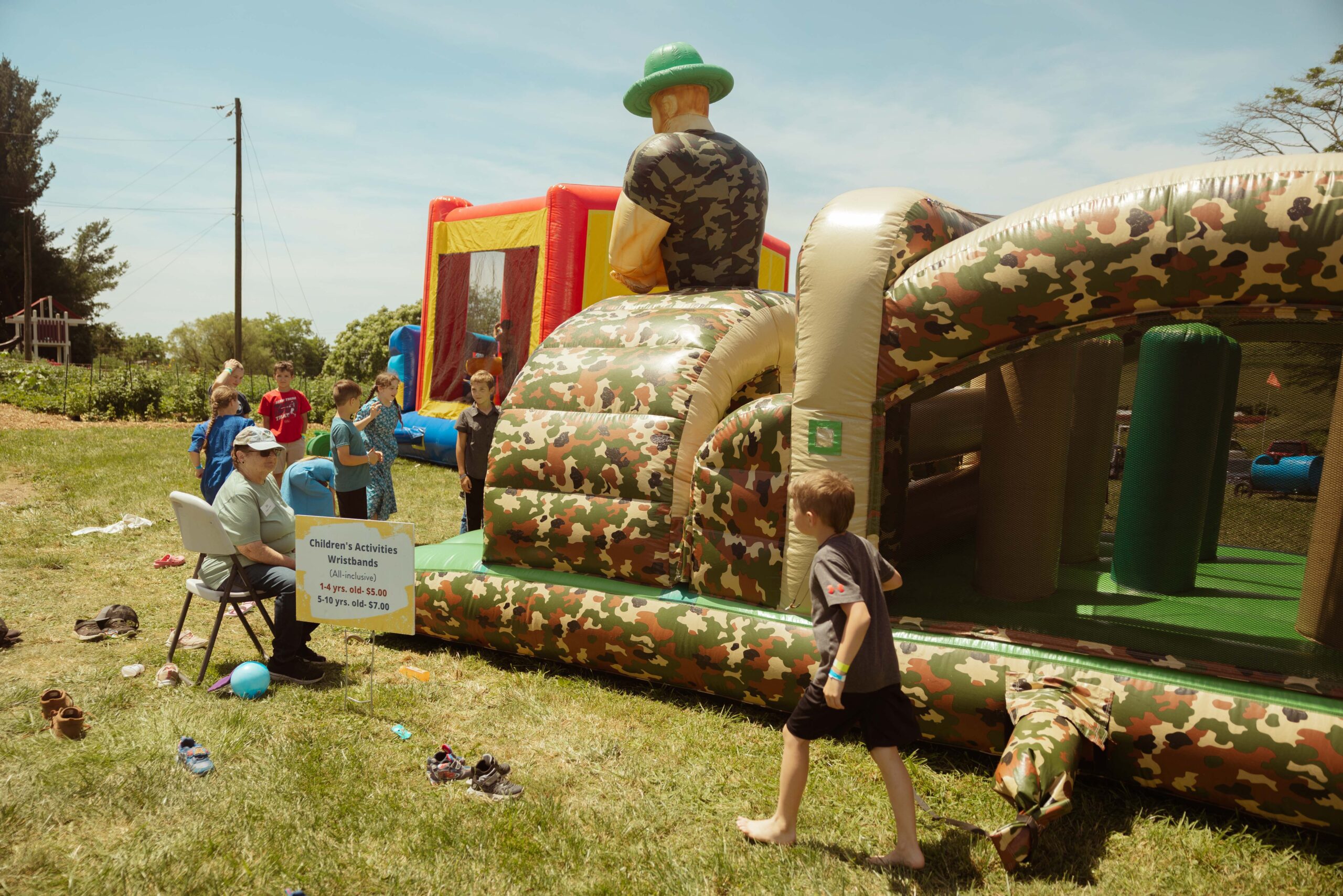 Children enjoying outdoor activities at a bounce house event, featuring a camouflage-themed inflatable structure. A sign indicates wristband pricing for children's activities, with various kids engaging in fun. The scene is set in a grassy area under a clear sky.