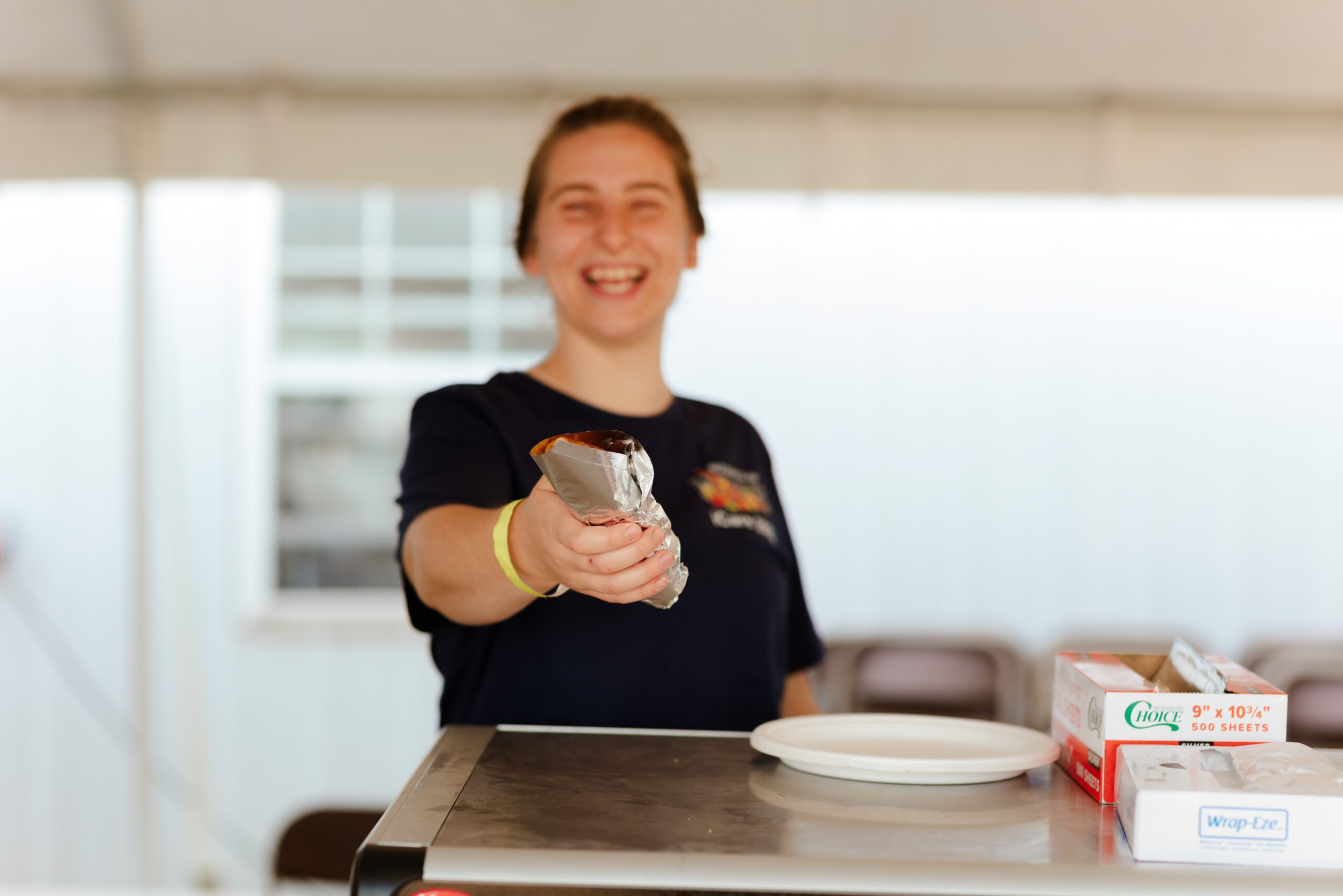 Smiling vendor handing out a foil-wrapped food item at an outdoor event with a table in the foreground and a white tent in the background.