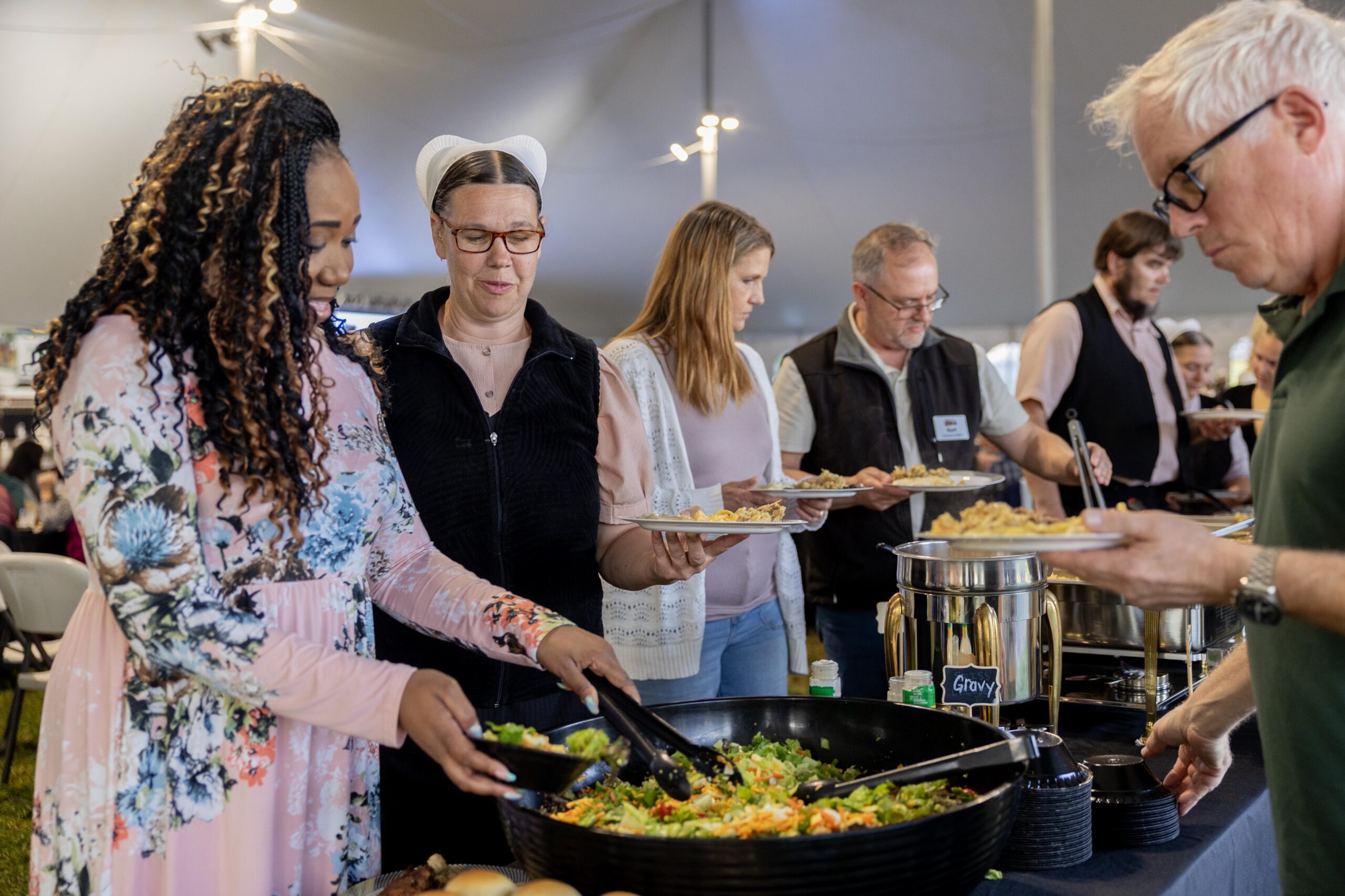 Community members enjoying a meal at a catered event, with a diverse group of individuals serving and selecting food from a large salad bowl and various dishes under a spacious tent.