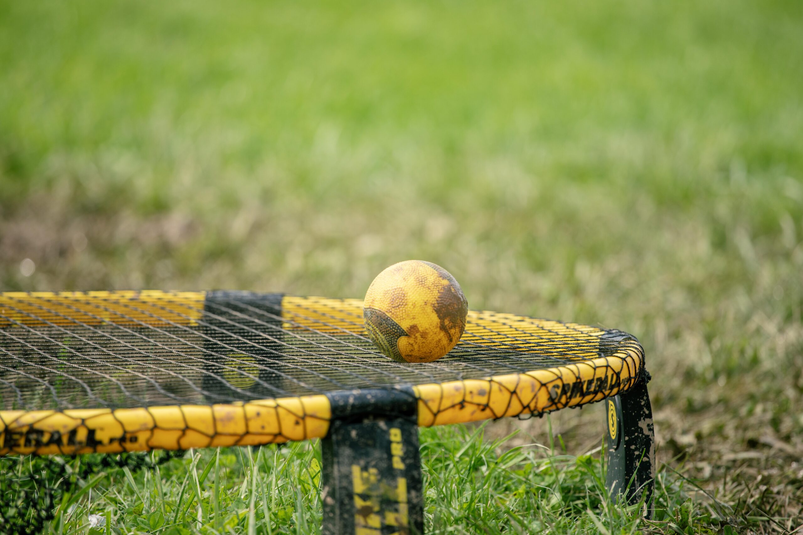 Close-up of a yellow ball resting on a yellow and black rebound net, set on a grassy field, showcasing equipment used for outdoor games and activities.