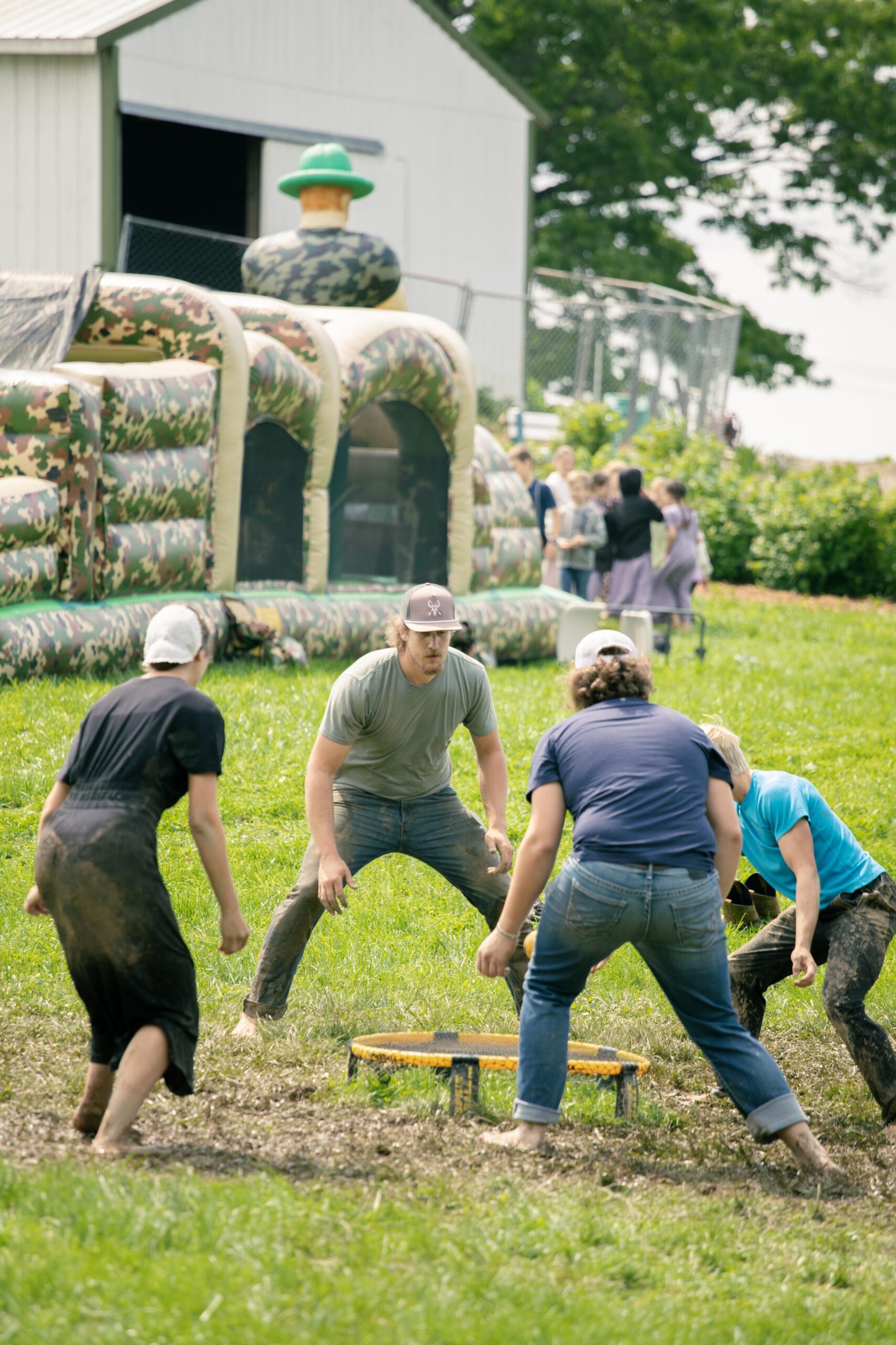 Group of young adults playing a muddy outdoor game on a grassy field, with a camouflage bouncy castle in the background, showcasing a fun and active summer event.