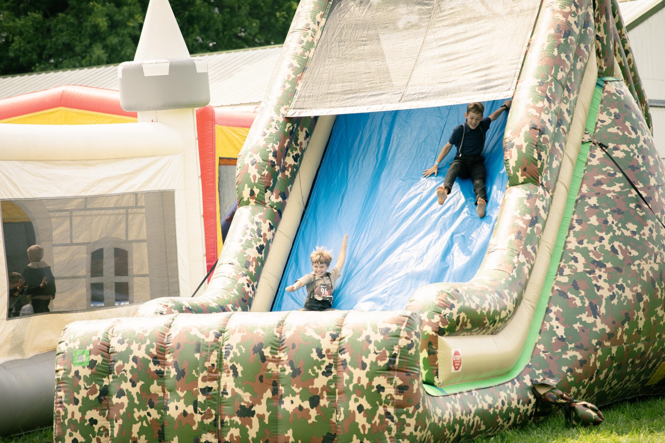 Kids playing on a large inflatable slide at a summer outdoor event, with one child sliding down enthusiastically while another prepares to slide, surrounded by colorful bounce houses in the background.