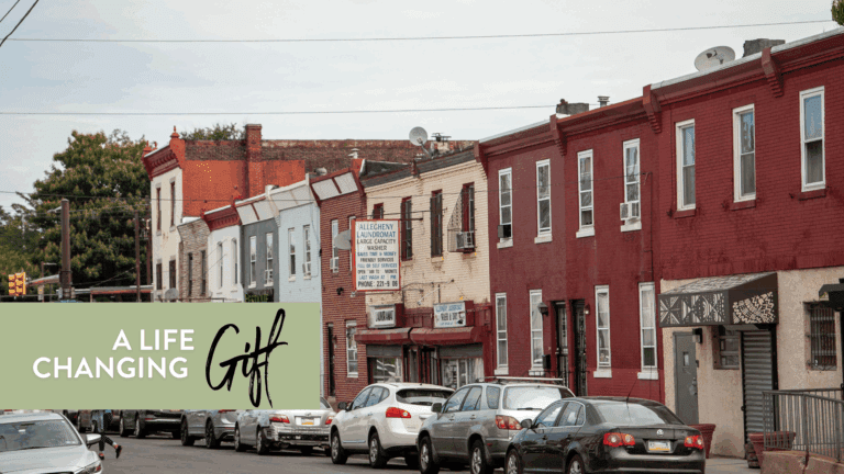Street view of a residential neighborhood featuring colorful brick buildings and parked cars, with a prominent sign for "Allegheny Laundromat." The scene captures a typical urban environment with greenery in the background. Text overlay reads "A Life Changing Gift."