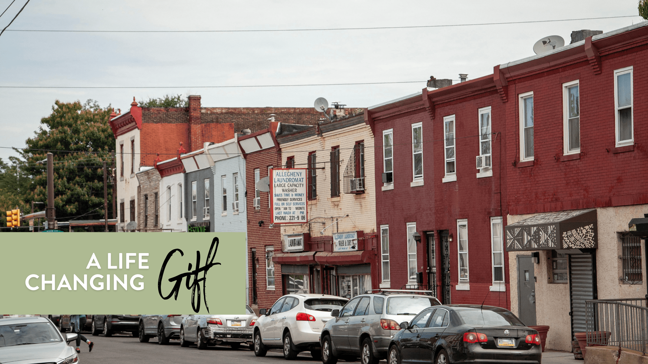 Street view of a residential neighborhood featuring colorful brick buildings and parked cars, with a prominent sign for "Allegheny Laundromat." The scene captures a typical urban environment with greenery in the background. Text overlay reads "A Life Changing Gift."