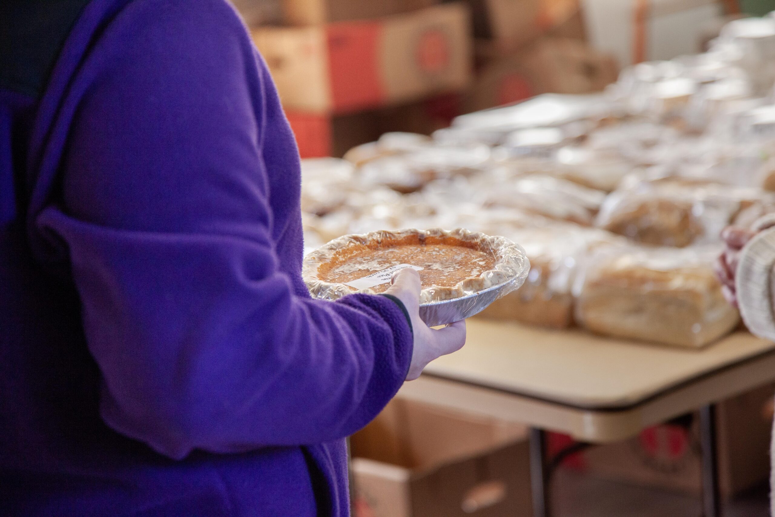 Person holding a freshly baked pumpkin pie in a silver pie pan at a food distribution event, with various baked goods visible on a table in the background.