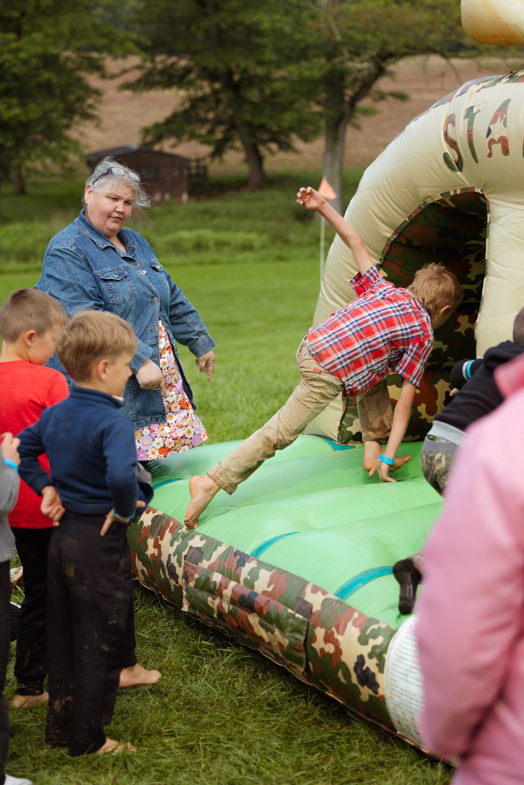 Children playing on a camouflage-themed inflatable bounce house, with one child jumping in while an adult supervises. The scene captures a fun outdoor activity, surrounded by green grass and trees, showcasing the excitement of kids at a community event or party.